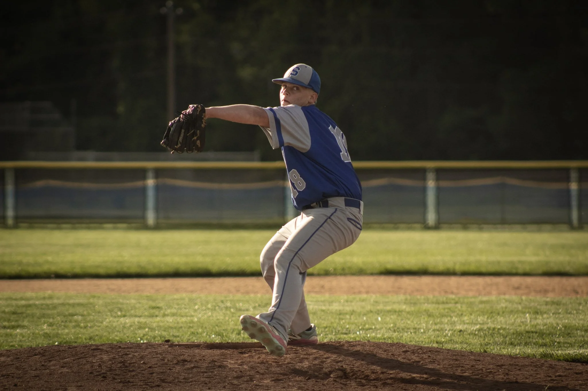 A young baseball pitcher in a blue and gray uniform pitching a ball on a baseball field during sunset.