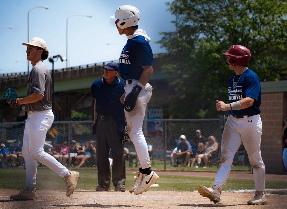 Young baseball players on a field, with one player jumping in the air, possibly celebrating, while others walk nearby. An umpire stands behind them and spectators sit in the background.