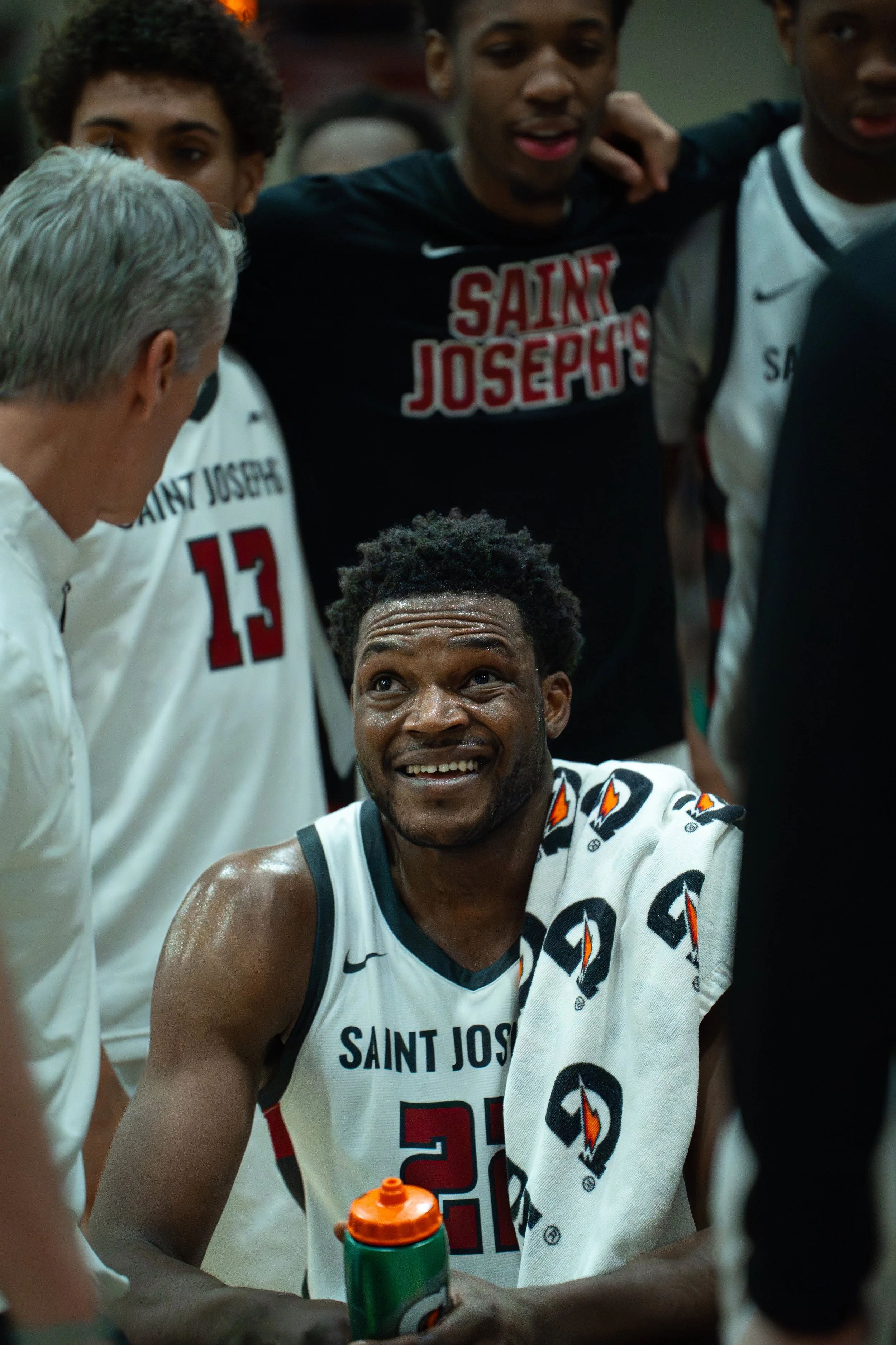 A group of basketball players and coach in a huddle during a timeout, with one player sitting, smiling, and holding a Gatorade bottle, wearing a white jersey with red and black accents, in a gymnasium.