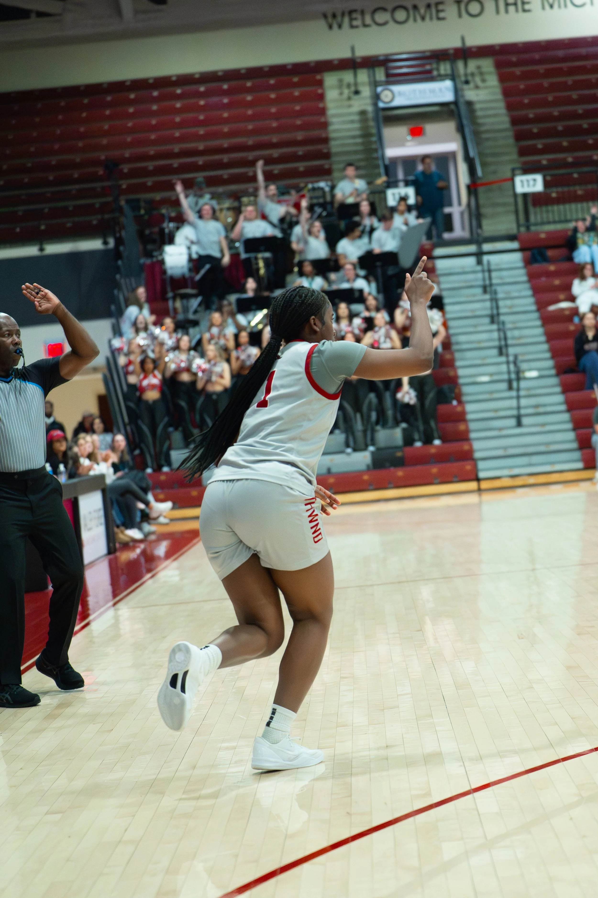 A female basketball player in a white uniform with red trim is running on the court, pointing upward with one finger, during a game in a gymnasium with spectators and musicians in the background.