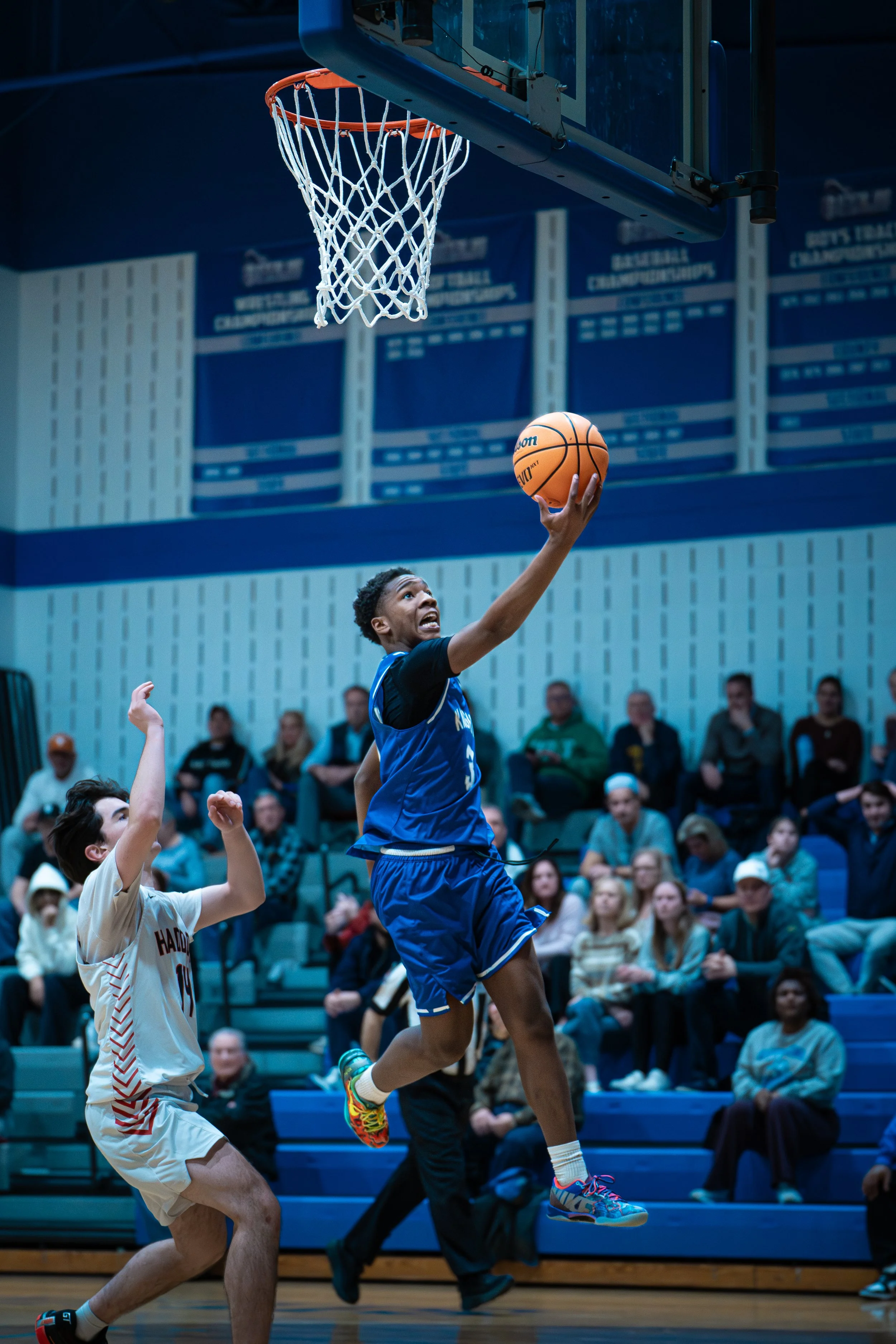 A young basketball player in a blue uniform leaps towards the basketball hoop, reaching to make a shot while another player in a beige uniform watches. Spectators are seated in the background.