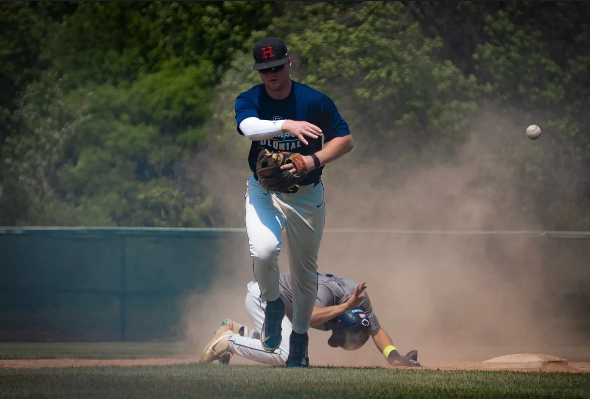 A baseball player running on the field while another player is on the ground in the background, dust rising from the dirt, with trees in the distance.