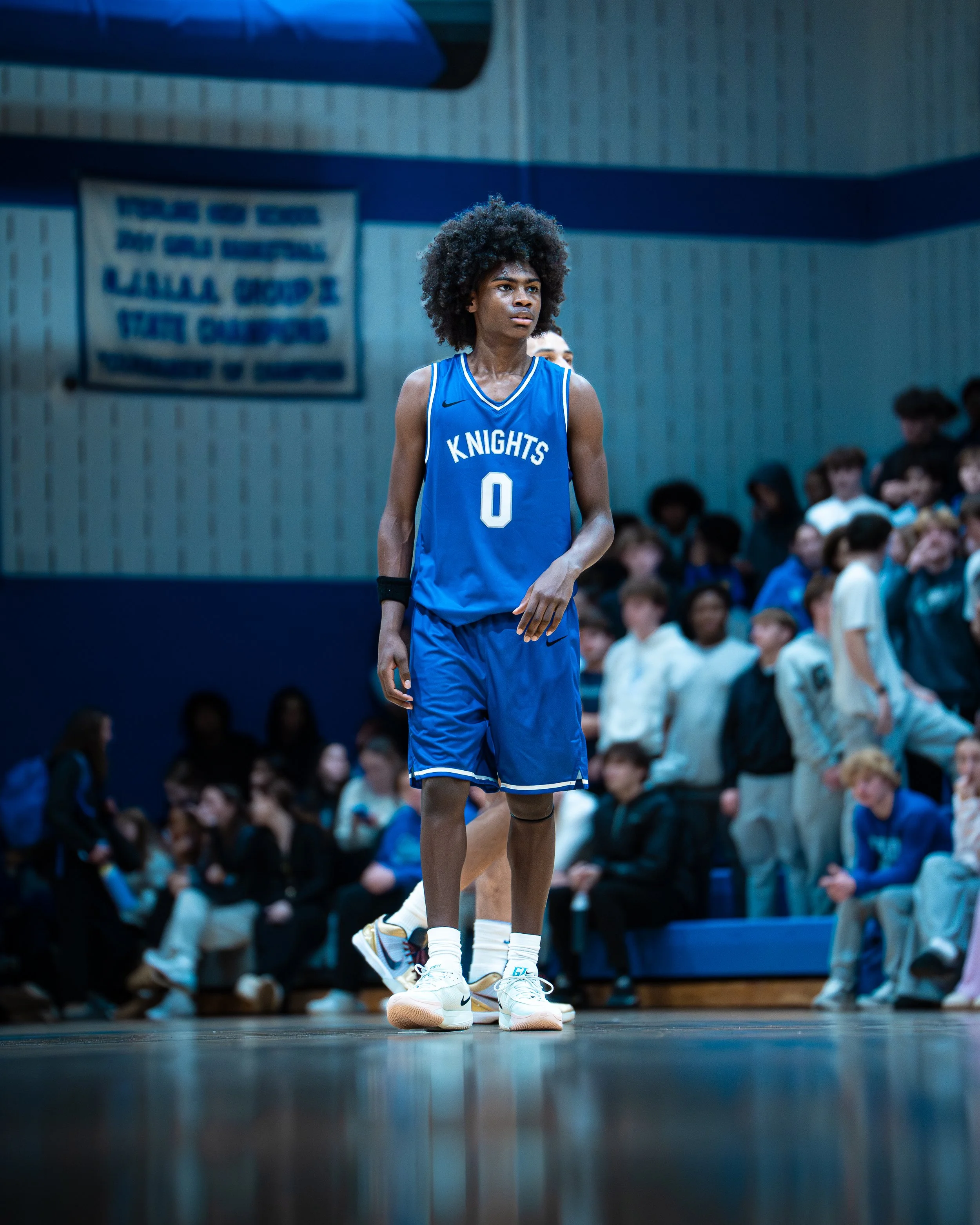 A basketball player with an afro hairstyle wearing a blue jersey with the word 'Knights' and the number 0 stands on a court during a game, with spectators in the background.