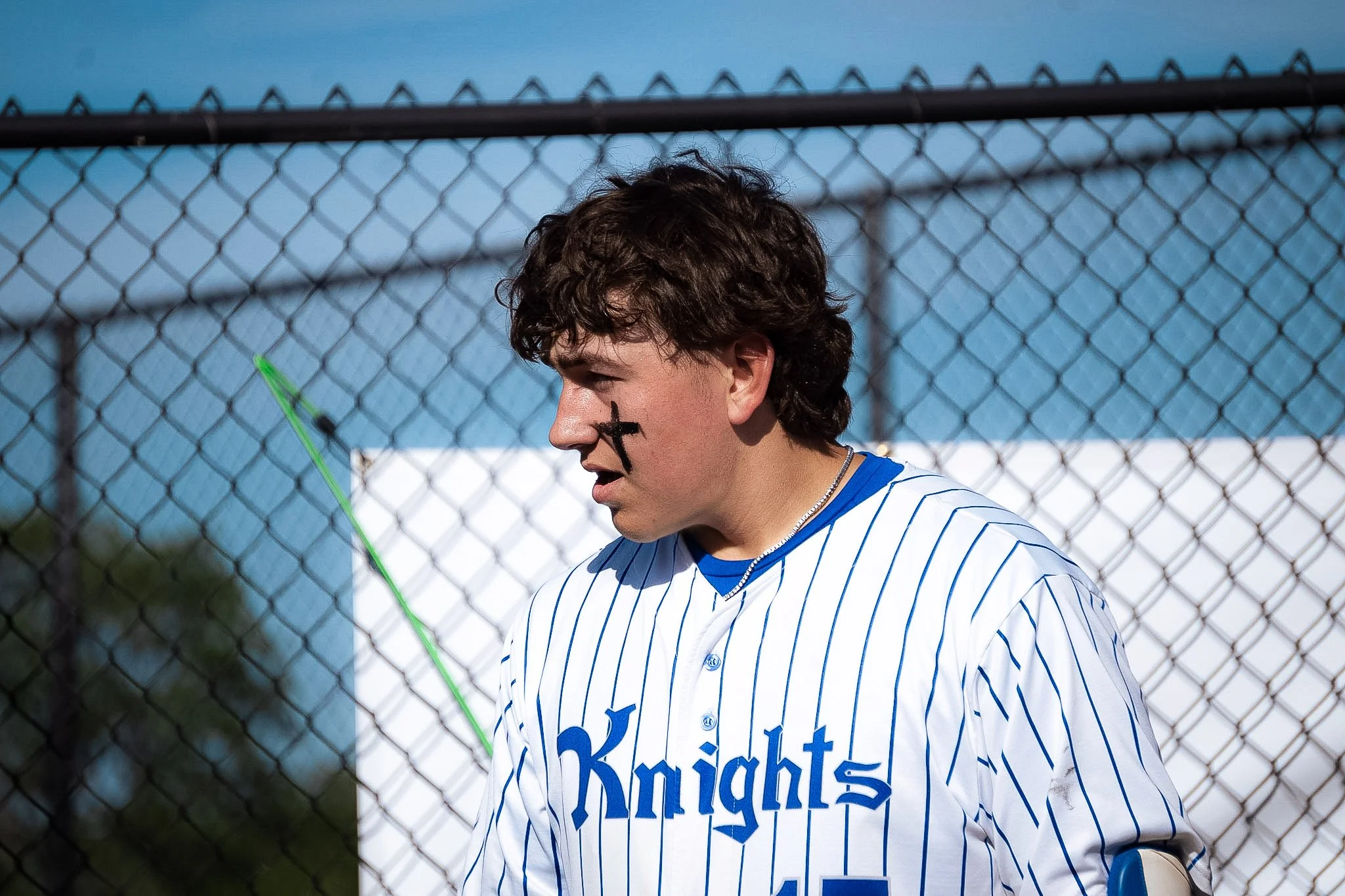 A teenage baseball player wearing a white jersey with blue pinstripes and the word 'Knights' on the front, standing near a chain-link fence during the daytime. He has dark, curly hair, a black eye-black under his eyes, and a black cross painted on hi