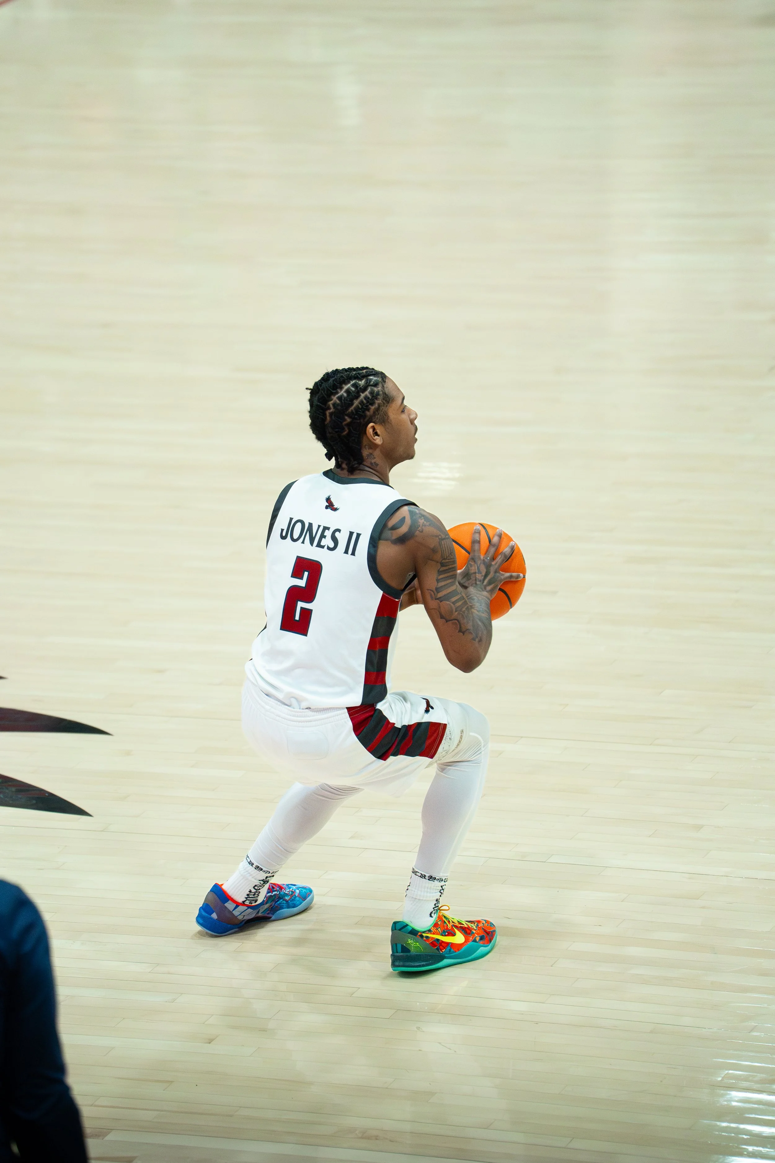 A basketball player in a white and black uniform with the number 2 and 'JONES II' on the back, crouching and holding a basketball, on a polished wooden court.