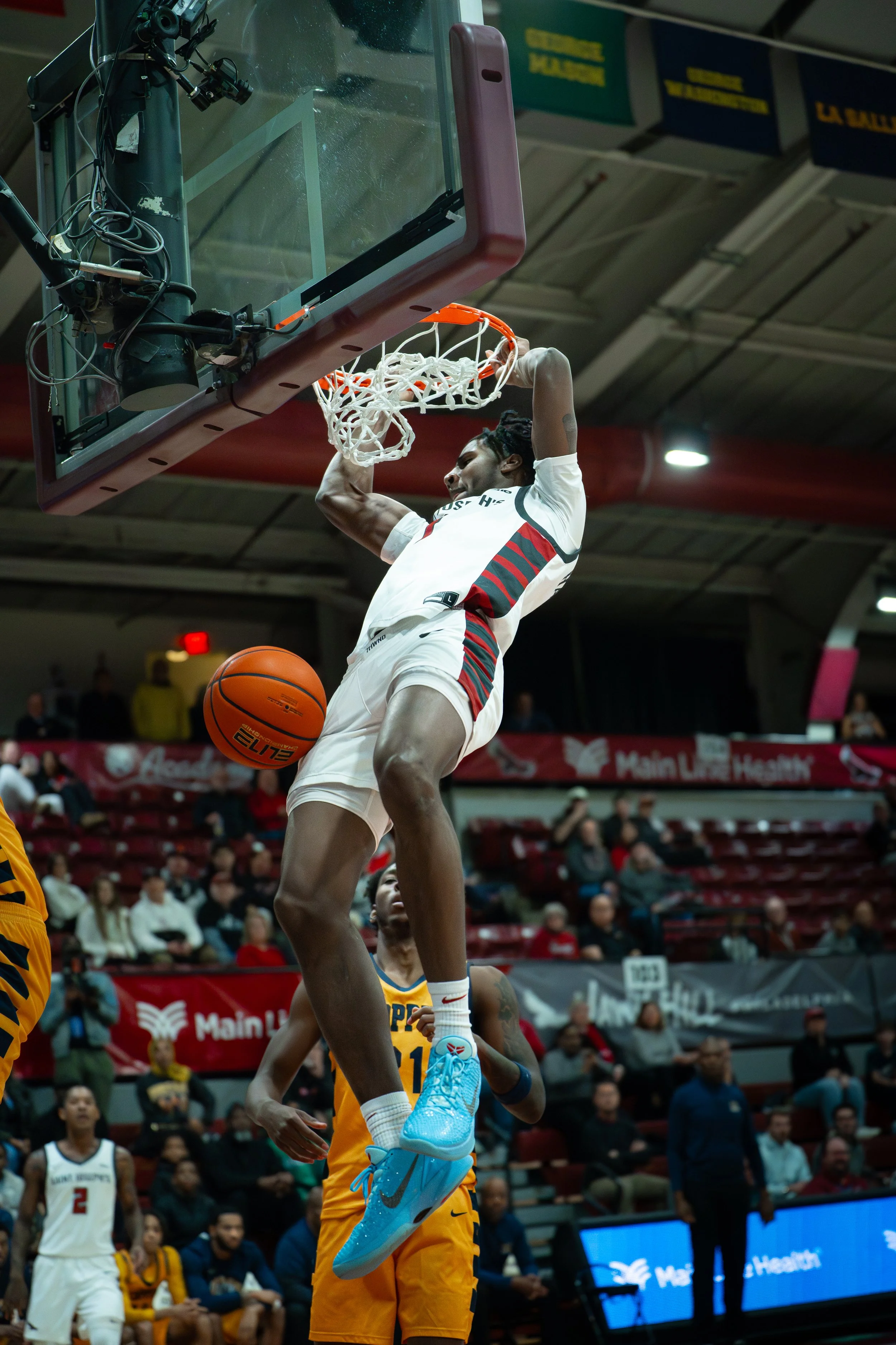 A basketball player in white and red uniform is dunking the ball through the hoop during a game. Opponent in yellow uniform is underneath, and spectators watch from the stands.