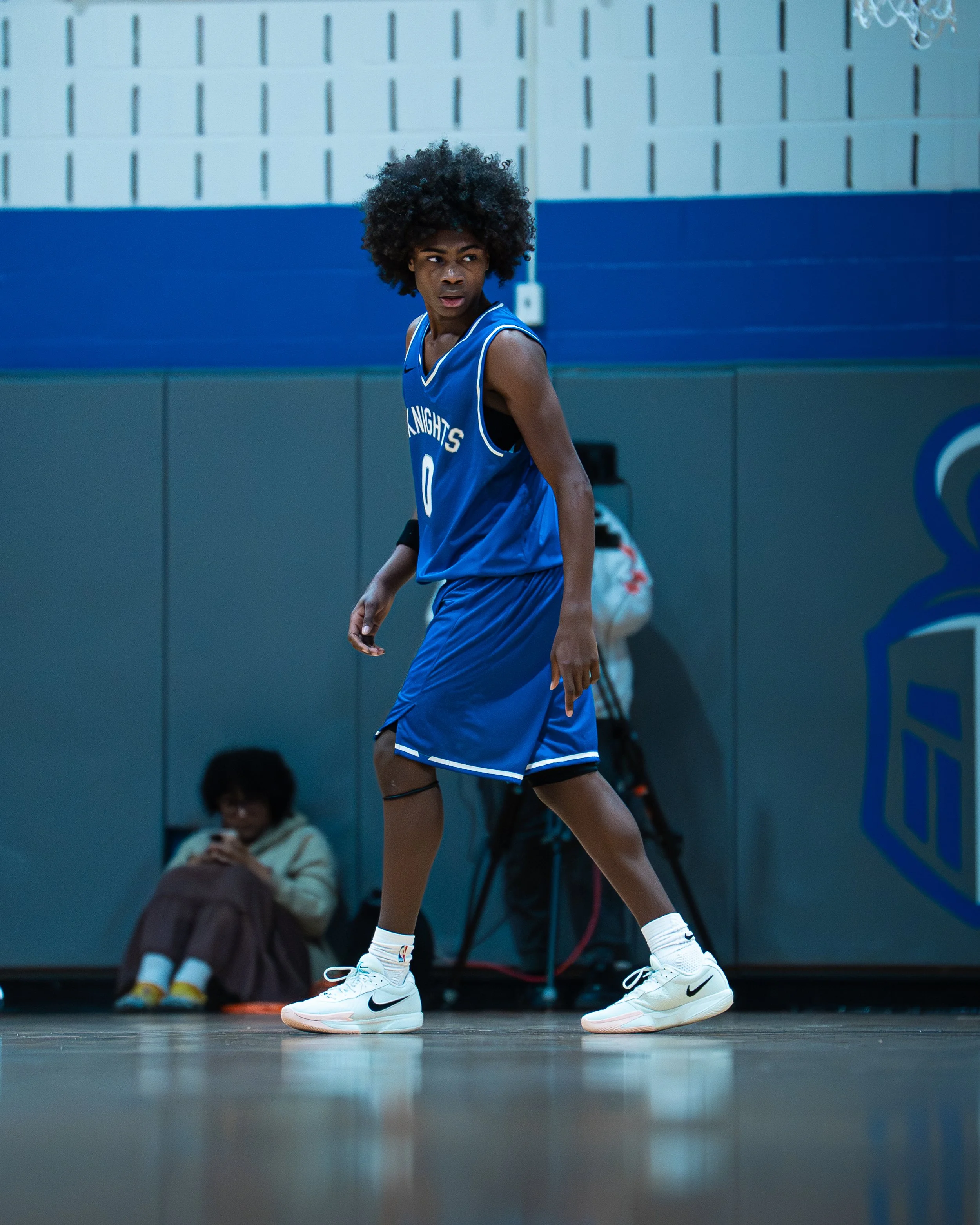 A young male basketball player in a blue jersey and shorts standing on a basketball court, looking to the side.