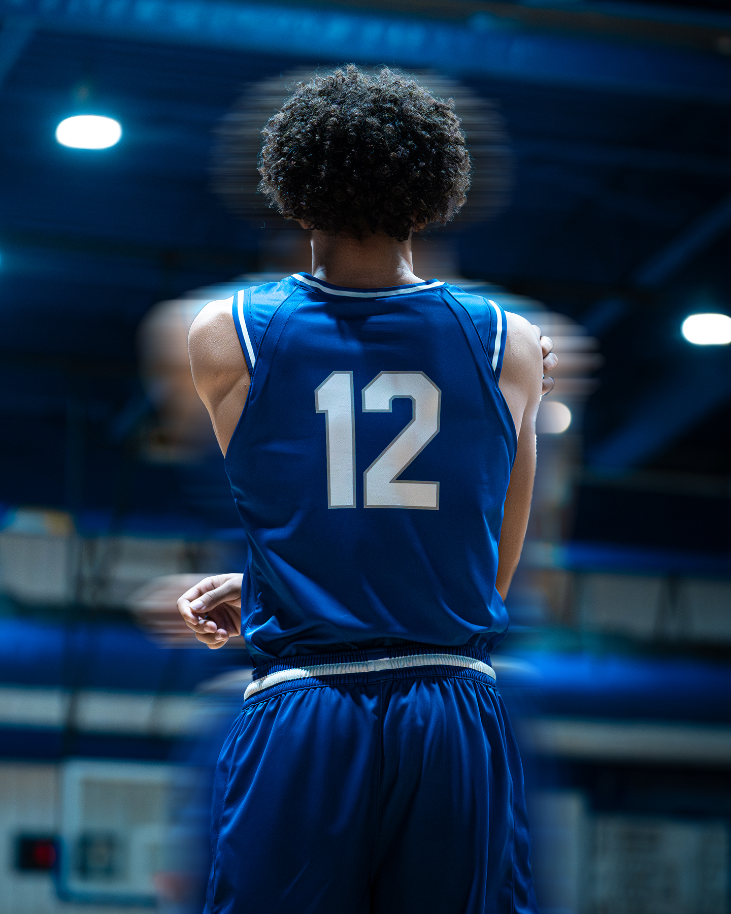 Back view of a basketball player in a blue jersey with the number 12 standing in an indoor basketball court.