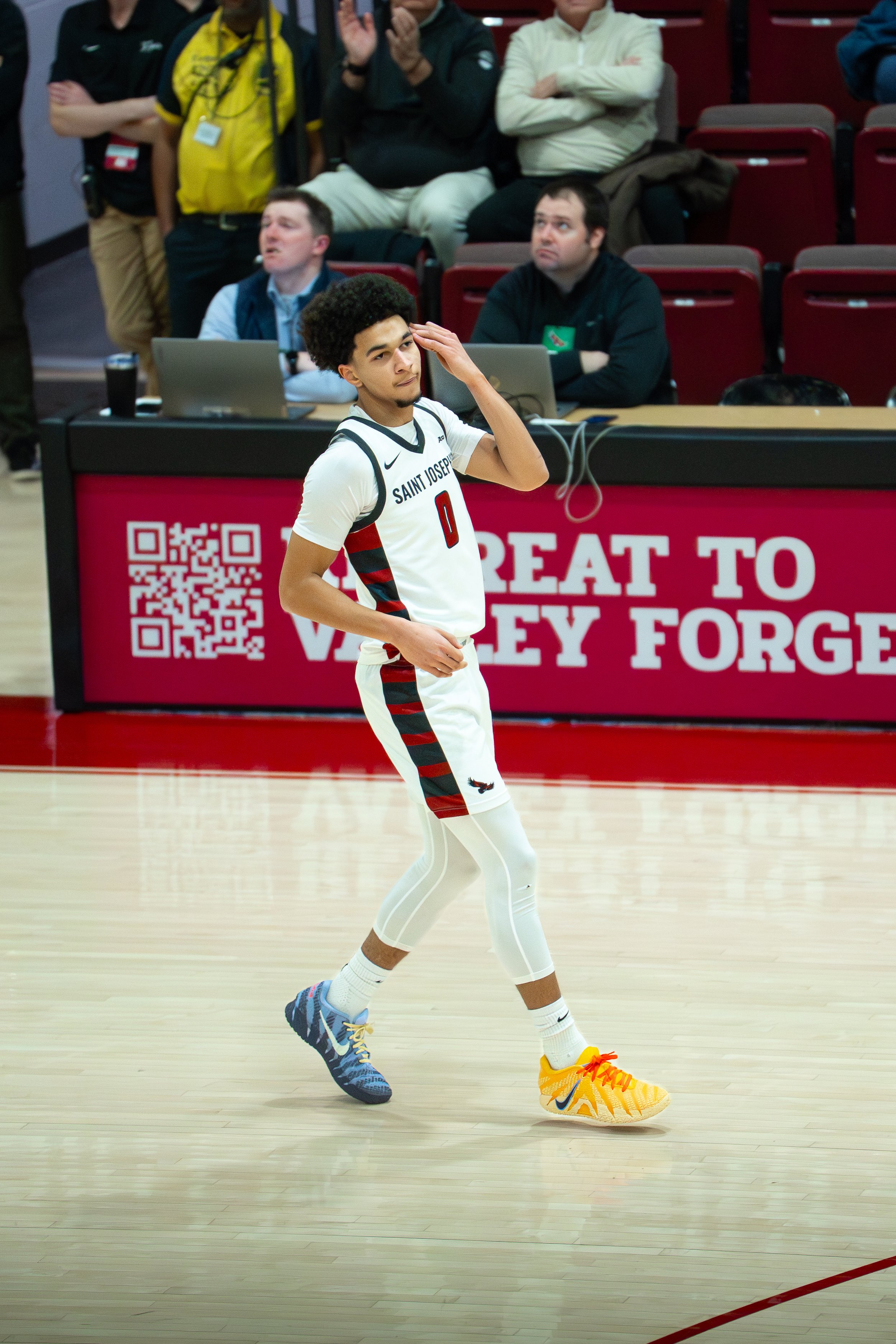 A young male basketball player in a Saint Joseph uniform walking on the court with a hand to his head, while spectators and officials watch from the stands and behind the scorer's table.