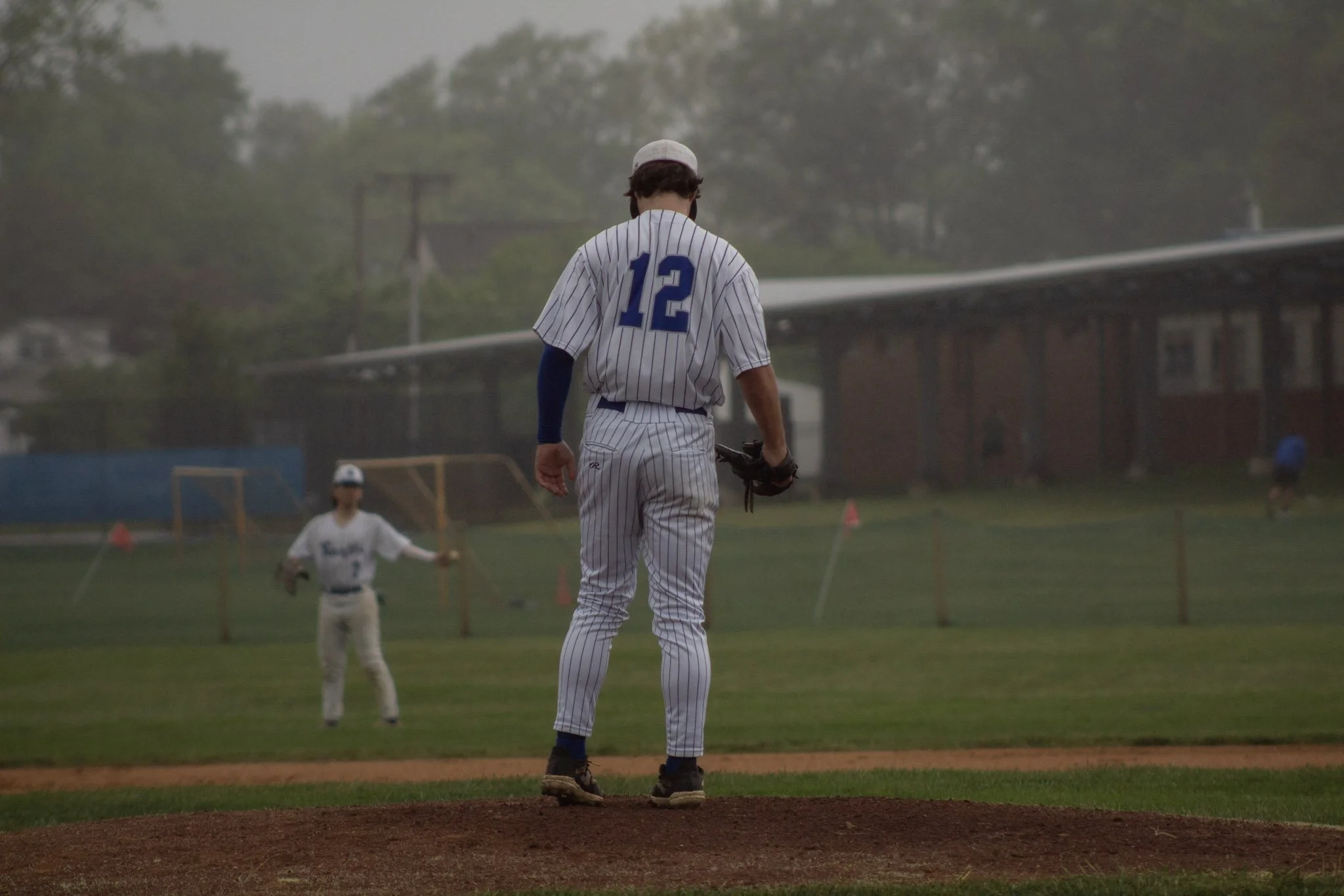 A baseball pitcher stands on the mound, facing the batter and the catcher in the background during a game in overcast weather.