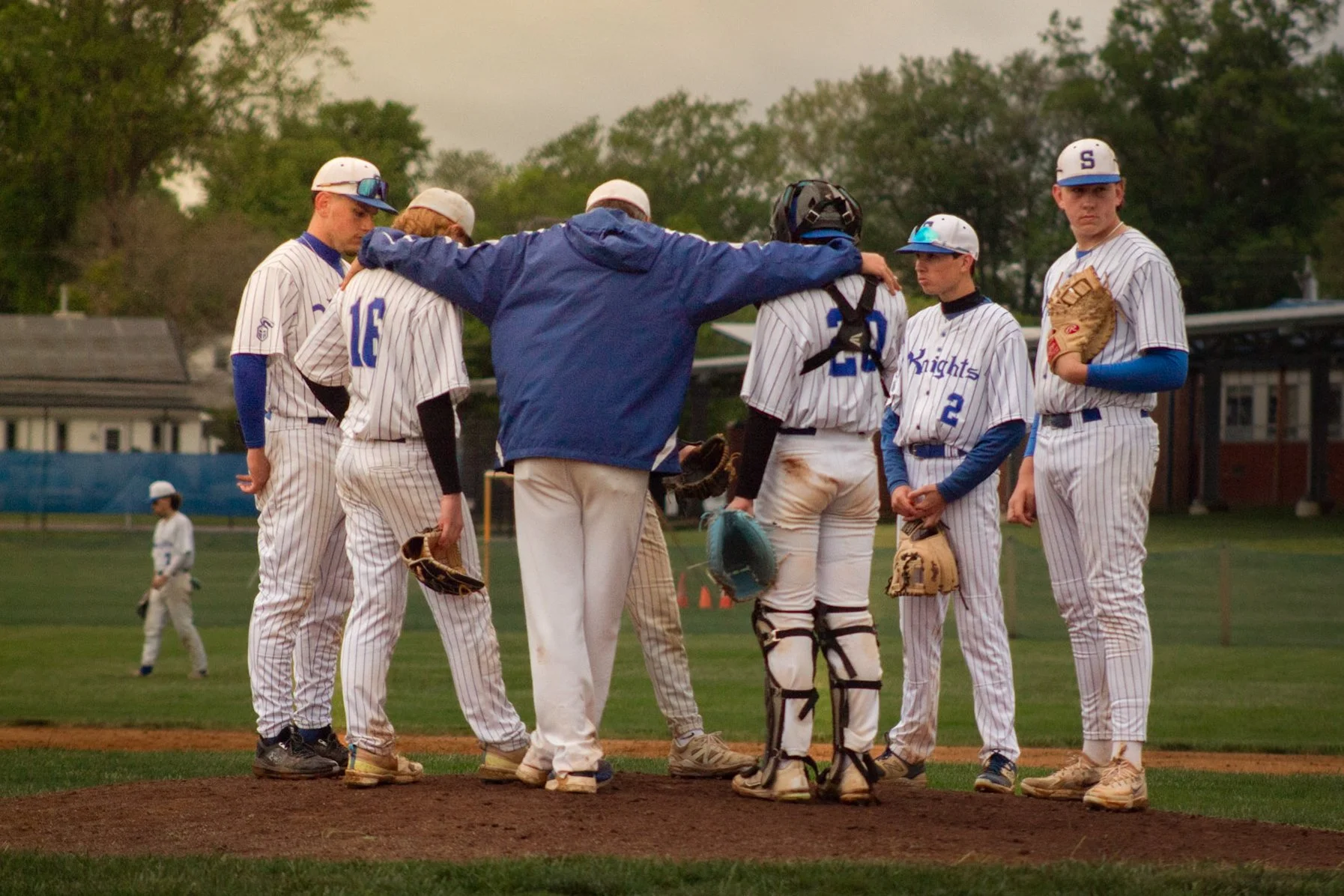 A group of young baseball players and coach standing on the pitcher's mound during a game, with a few infielders and outfielders in the background.
