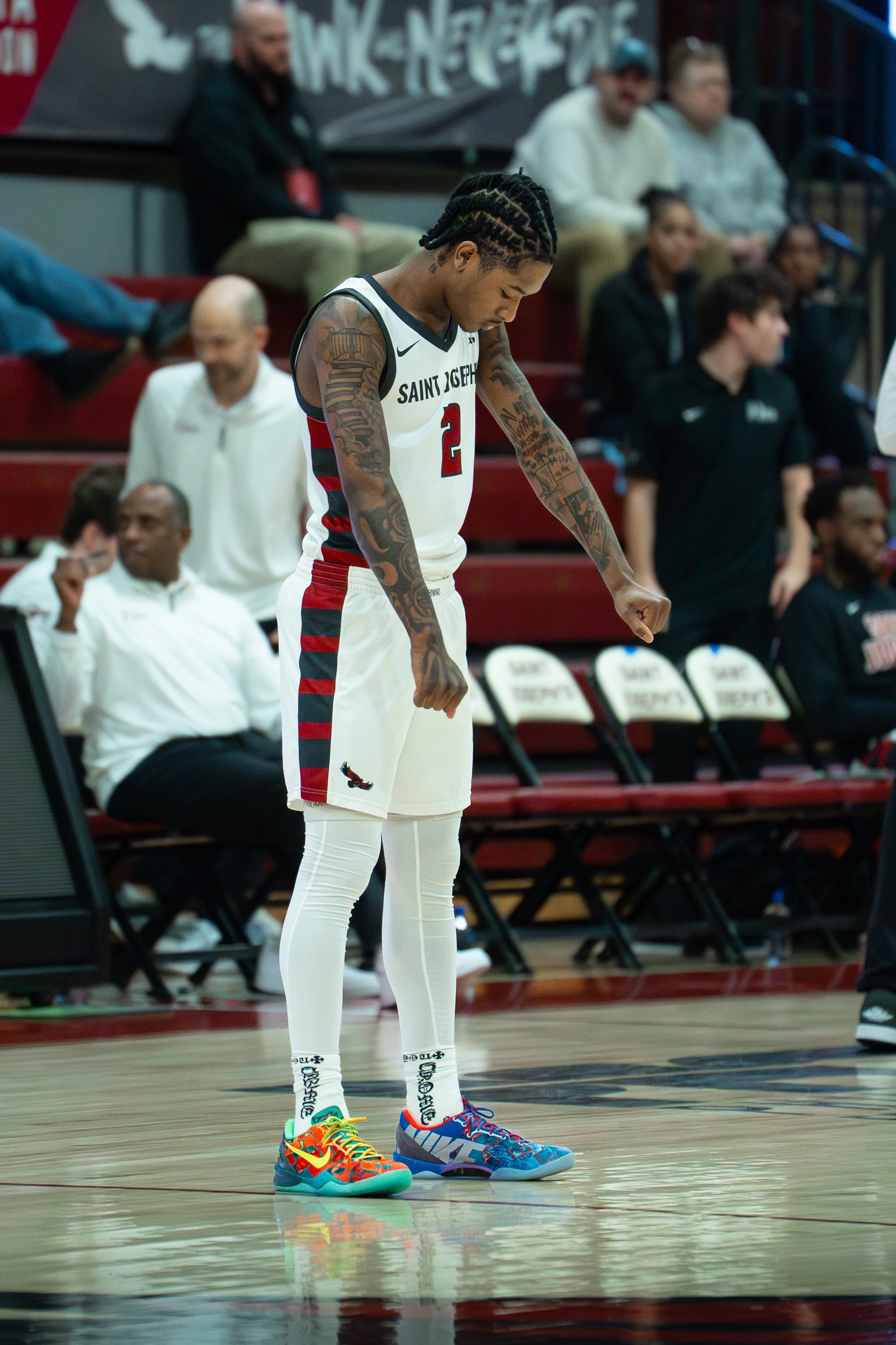 A basketball player wearing a Saint Joseph jersey with the number 2, colorful sneakers, and white leggings, standing on a basketball court with his head down. In the background, spectators and team staff are seated on benches.