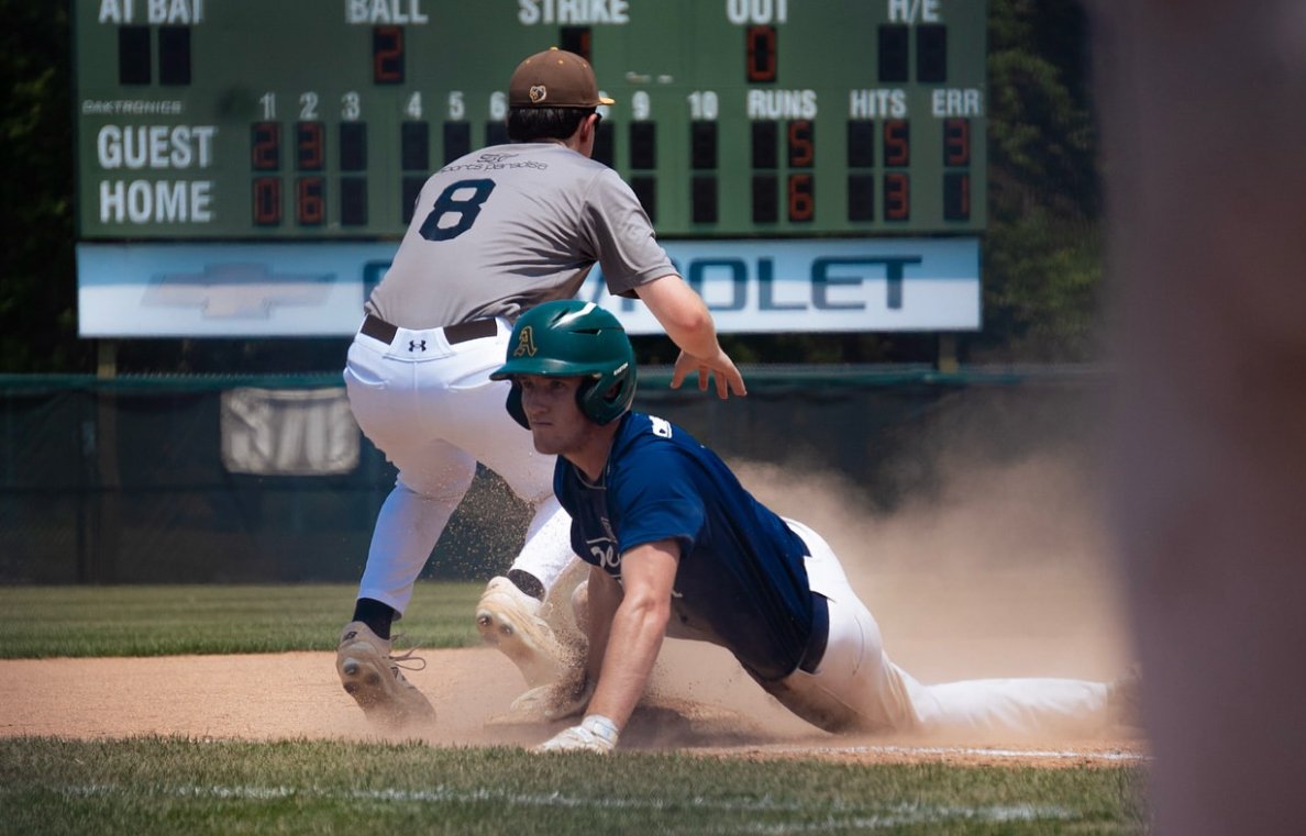 A baseball player in a blue uniform sliding into a base, while a player in a gray uniform attempts to tag him out. The scoreboard in the background shows game statistics.