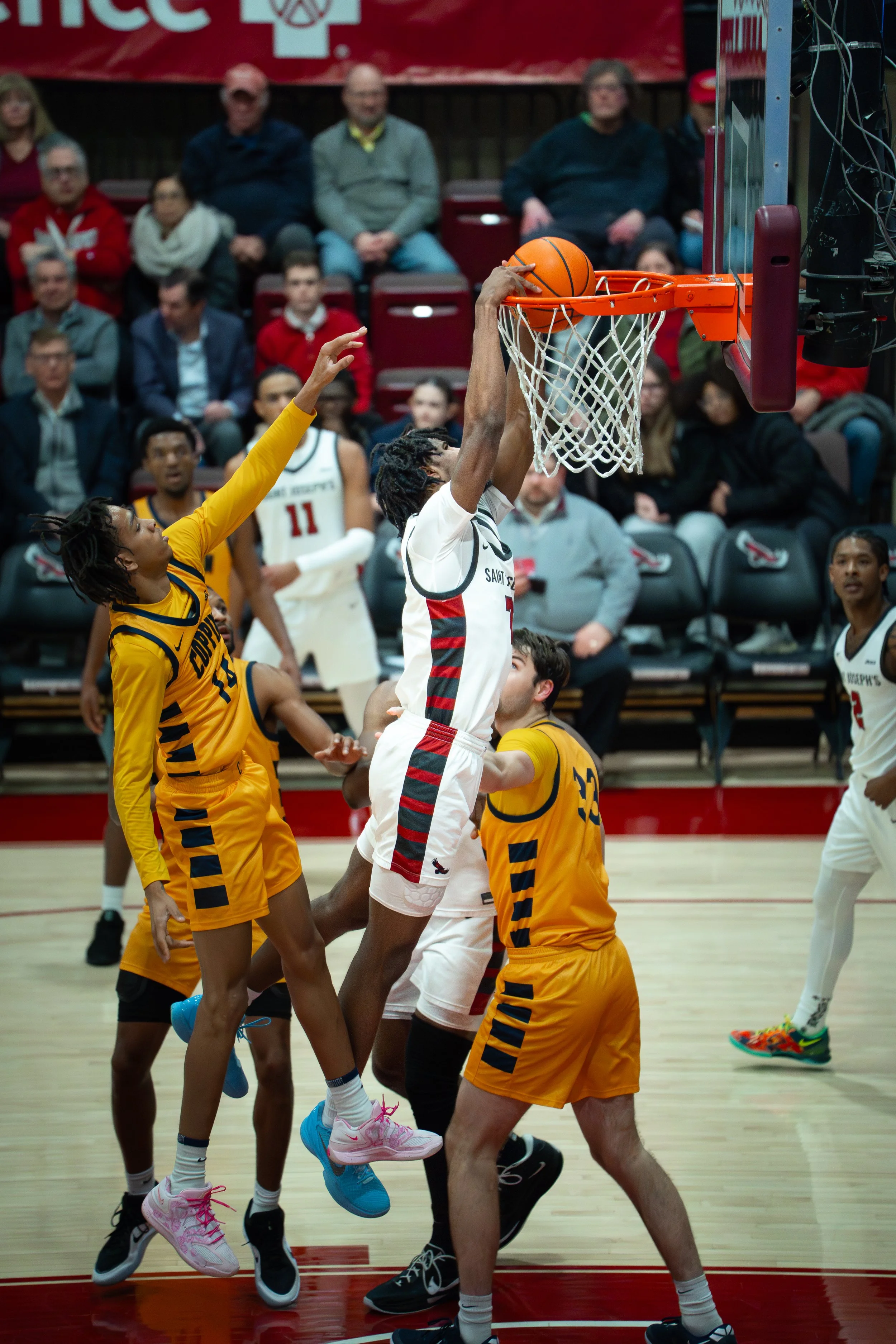 A basketball player in a white uniform is dunking the ball into the hoop while wearing a white jersey with black and red stripes and pink shoes. Two players in yellow uniforms are defending him, one reaching out and the other holding him. People are 