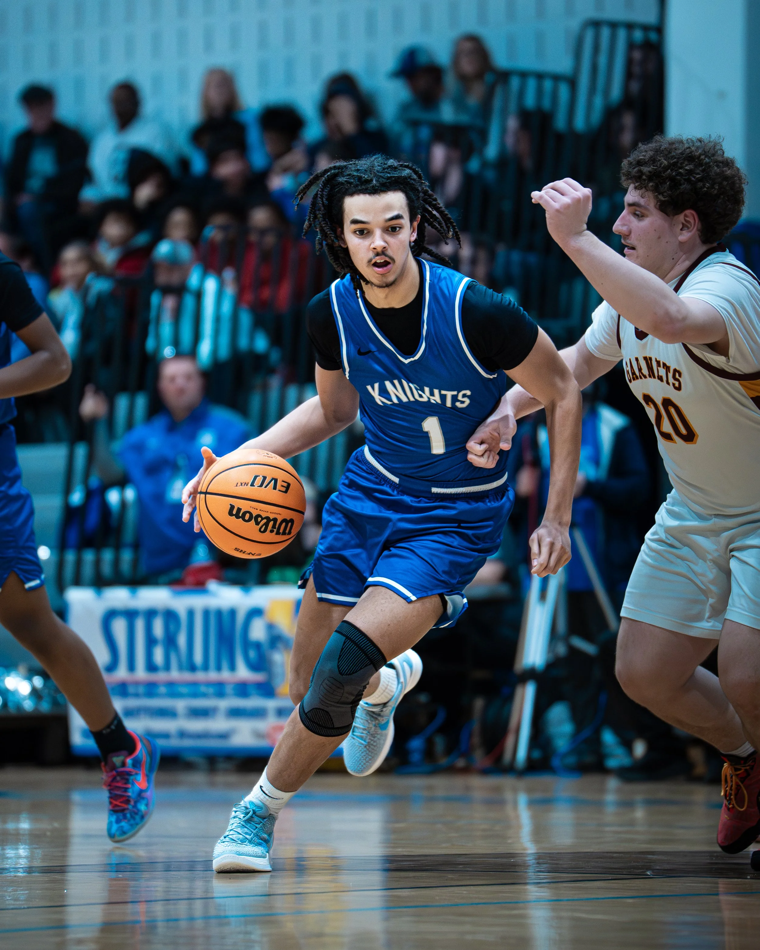 A young male basketball player with dreadlocks wearing a blue uniform with 'Knights' and the number 1, dribbling a basketball during a game. An opponent in a white and yellow uniform with the number 20 is defending him. Spectators are visible in the 