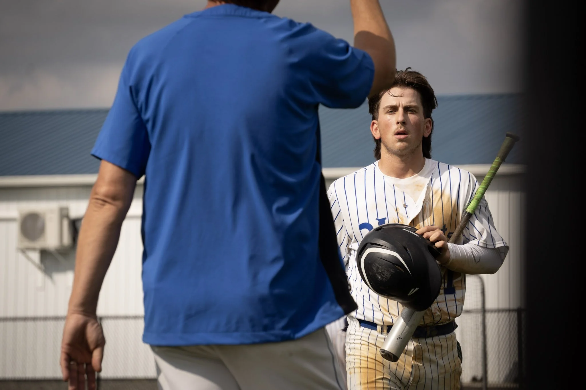 A young man in a baseball uniform holding a bat and helmet, standing in front of a coach, appears fatigued or disappointed after a game.