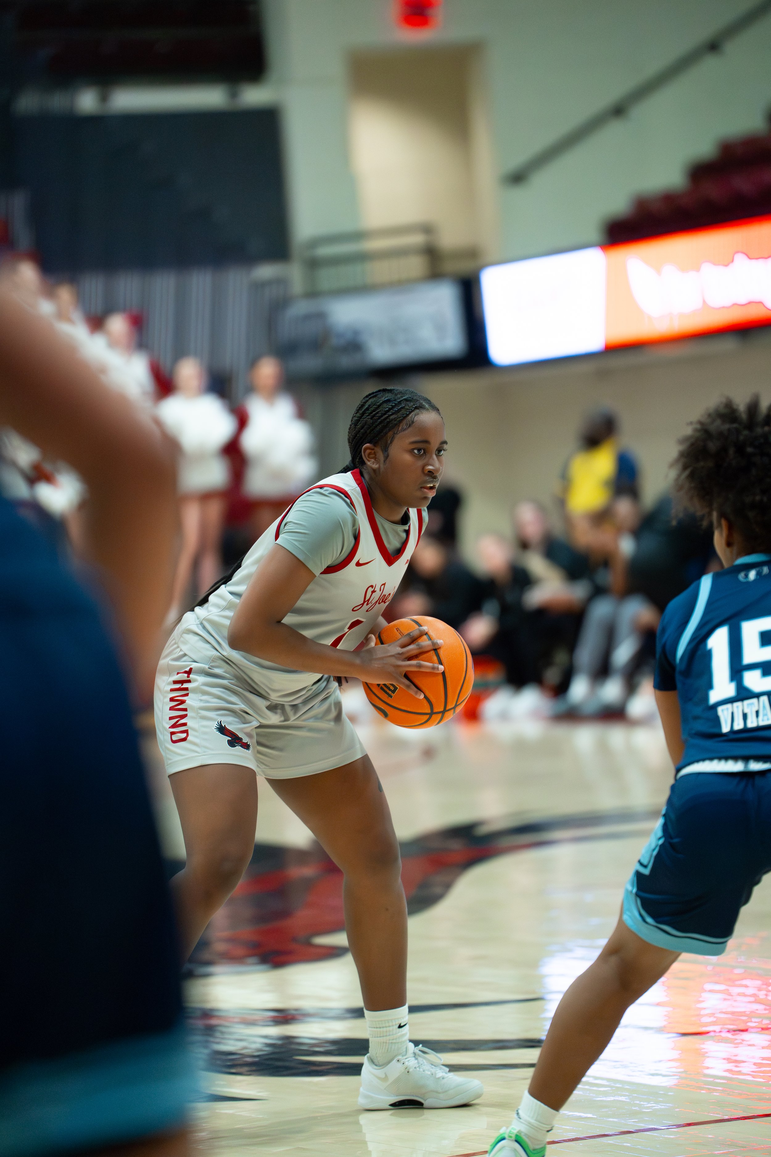 A female basketball player wearing a light gray and red jersey with 'ST. JOSEPH' written on it, holding a basketball on the court during a game. She is facing a defender wearing a navy blue jersey with the number 15.