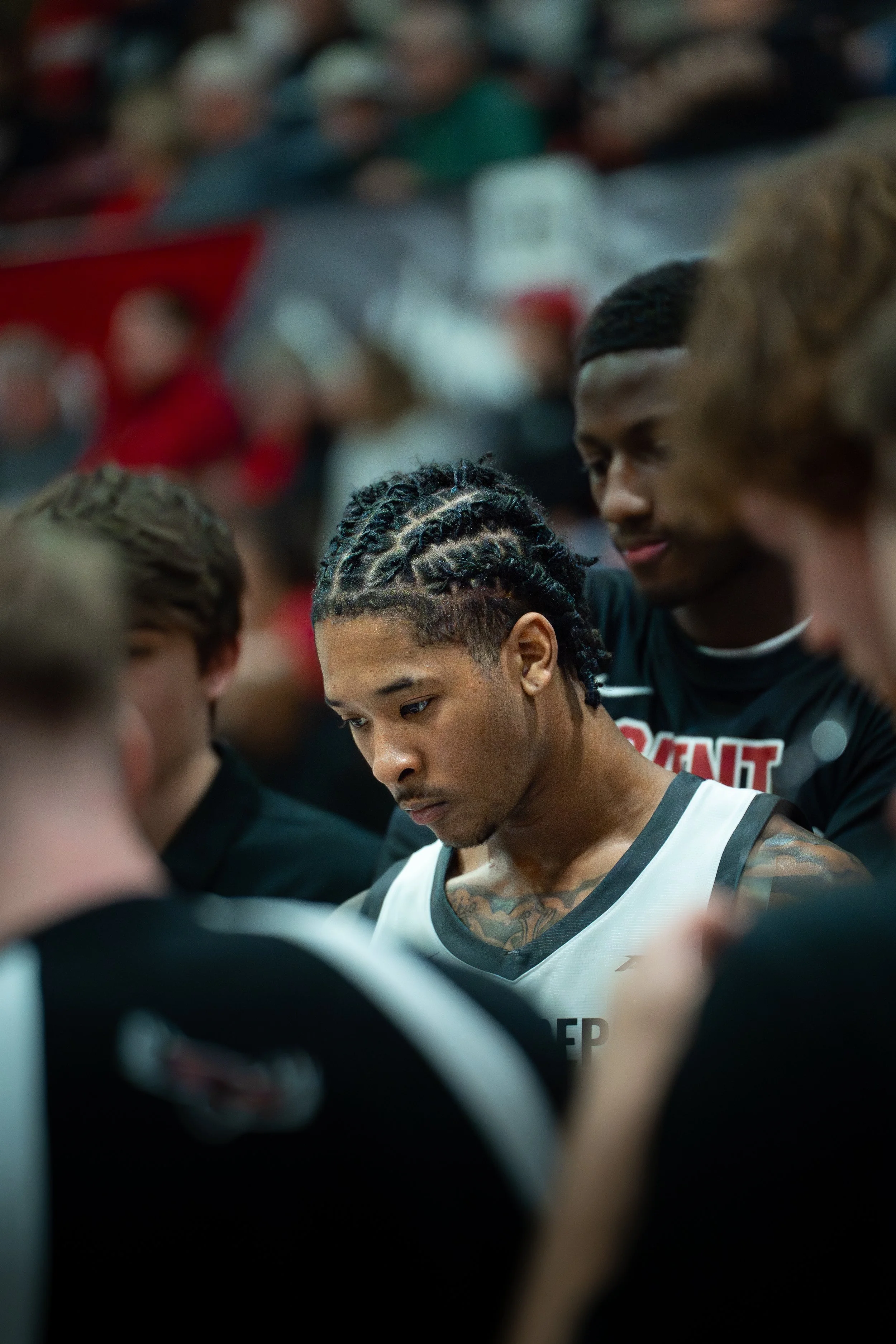 Young basketball player with braided hair and tattoos, standing with his teammates during a team huddle.