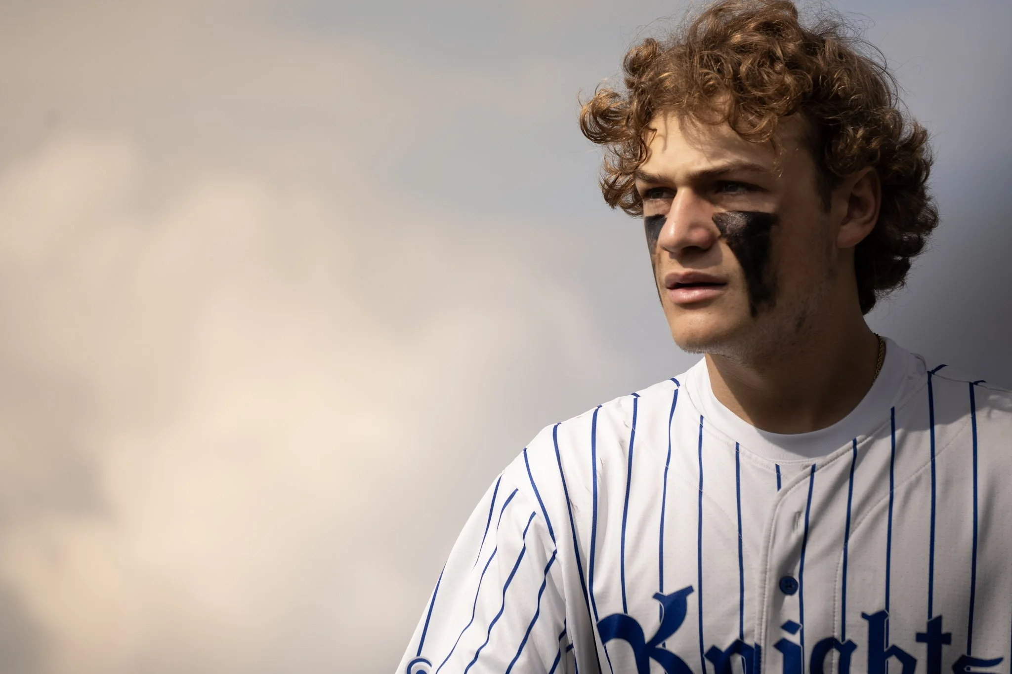 Young man with curly hair wearing a white baseball jersey with blue pinstripes and black face paint, looking off into the distance against a cloudy sky.