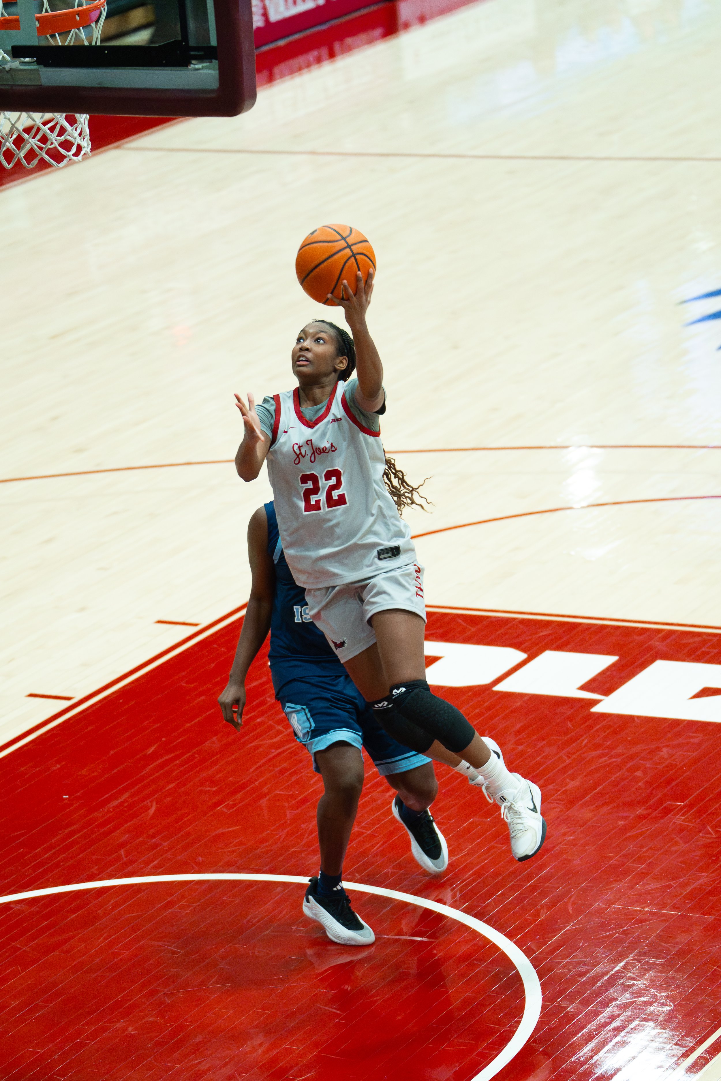 A female basketball player wearing a white jersey with the number 22 is jumping near the basket, holding a basketball in mid-air, about to make a shot. She is being defended by a player in a blue uniform. The court has red and white markings.