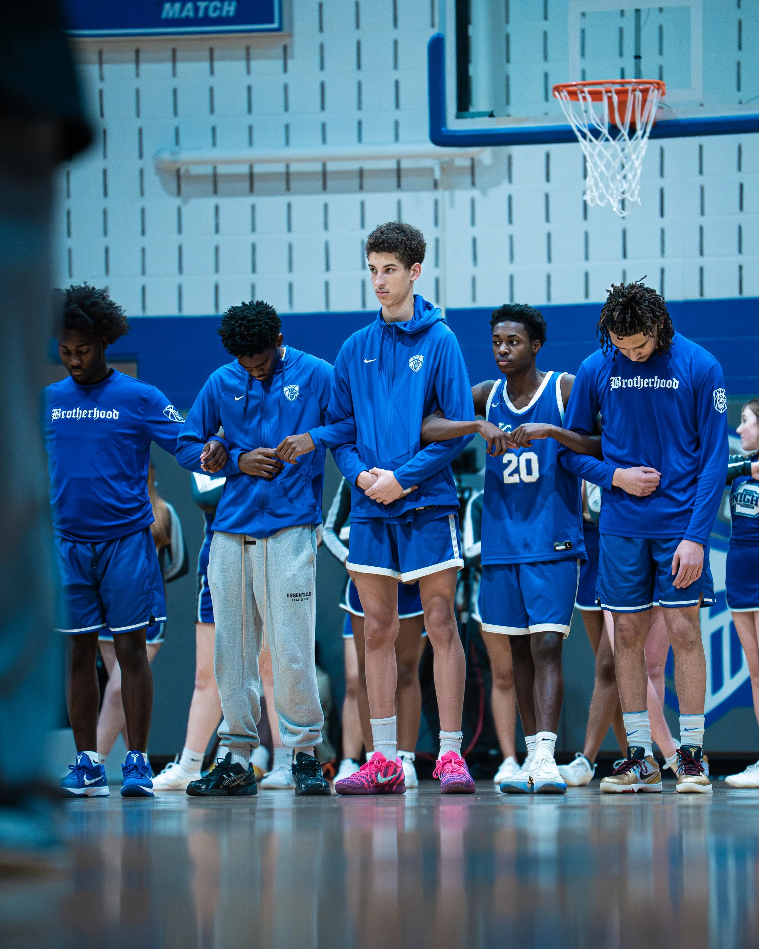 A group of young basketball players standing in a gym, holding hands and bowing their heads during a team moment.
