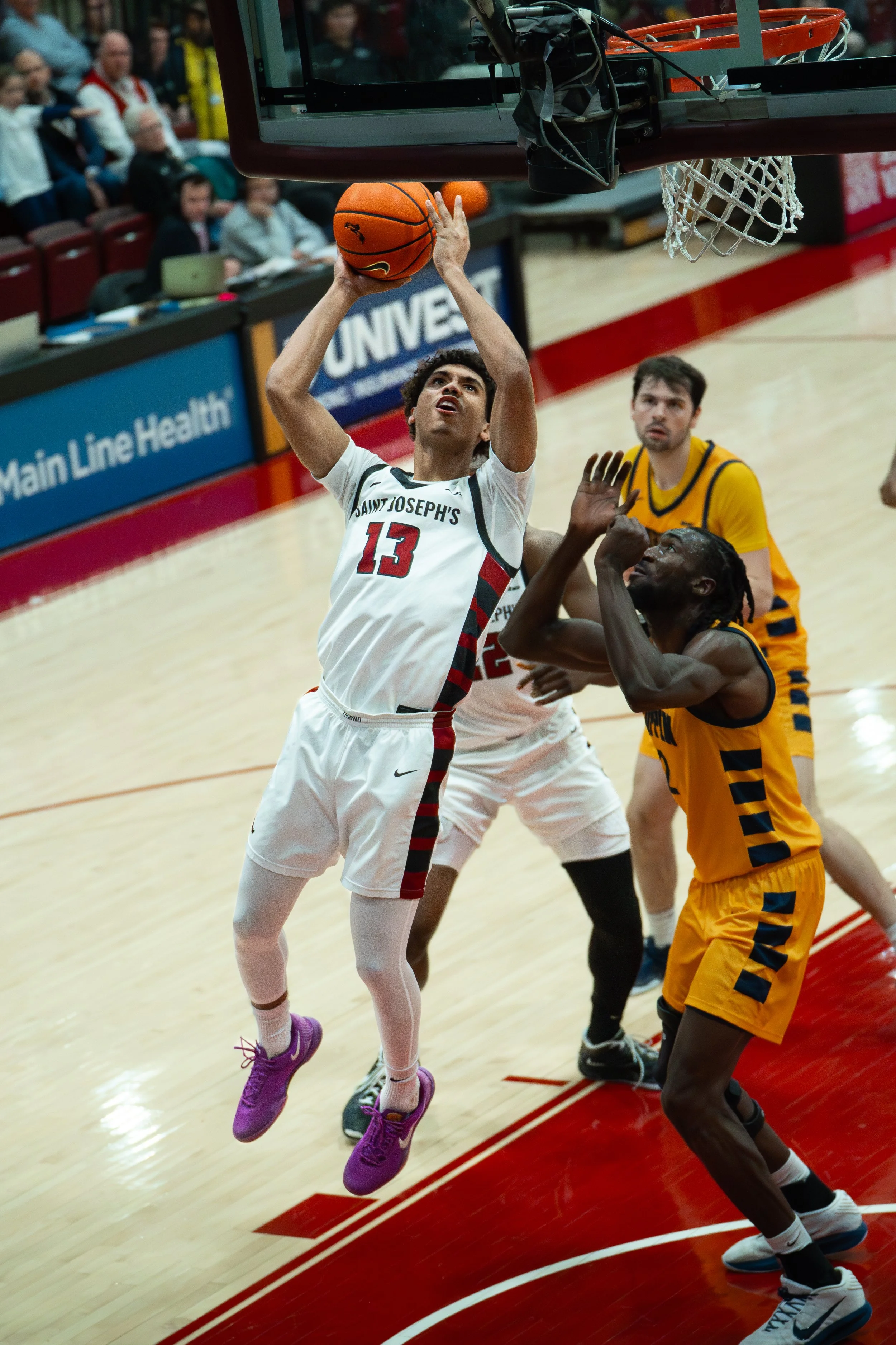 A basketball player in a white jersey with red and black accents, wearing number 13 and purple shoes, is jumping to make a shot near the hoop while an opponent in an orange jersey attempts to block him. Other players and spectators are visible in the
