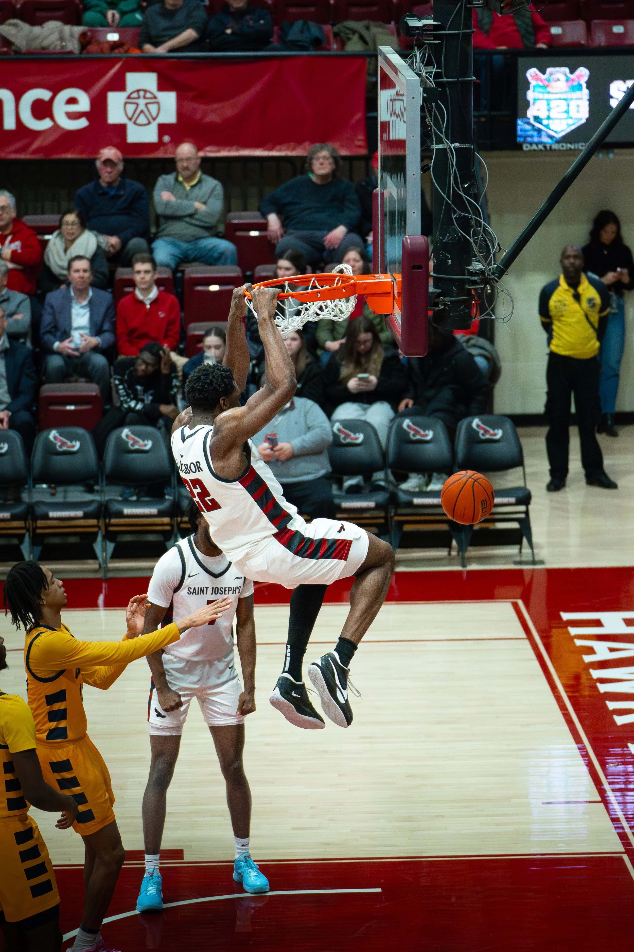 A basketball player in a white jersey with red and black accents jumps near the basket, holding the basketball, with spectators in the background.
