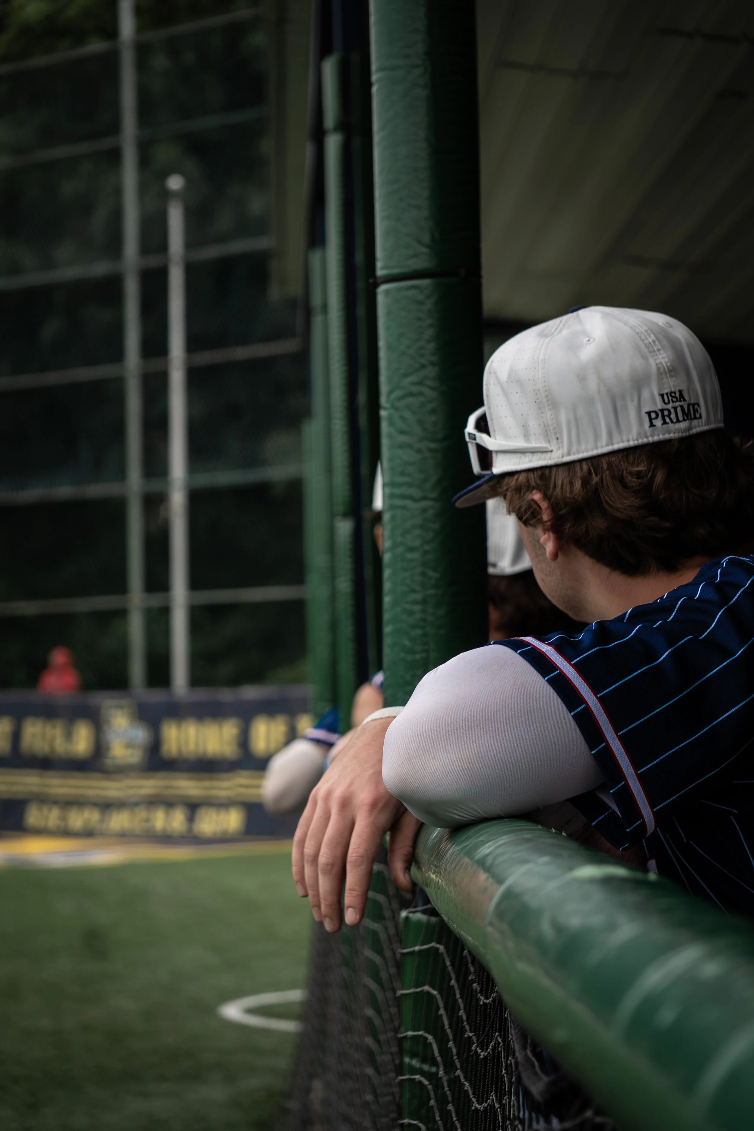 A baseball player in a blue jersey with red and white trim sitting in the dugout, leaning on a green metal railing.