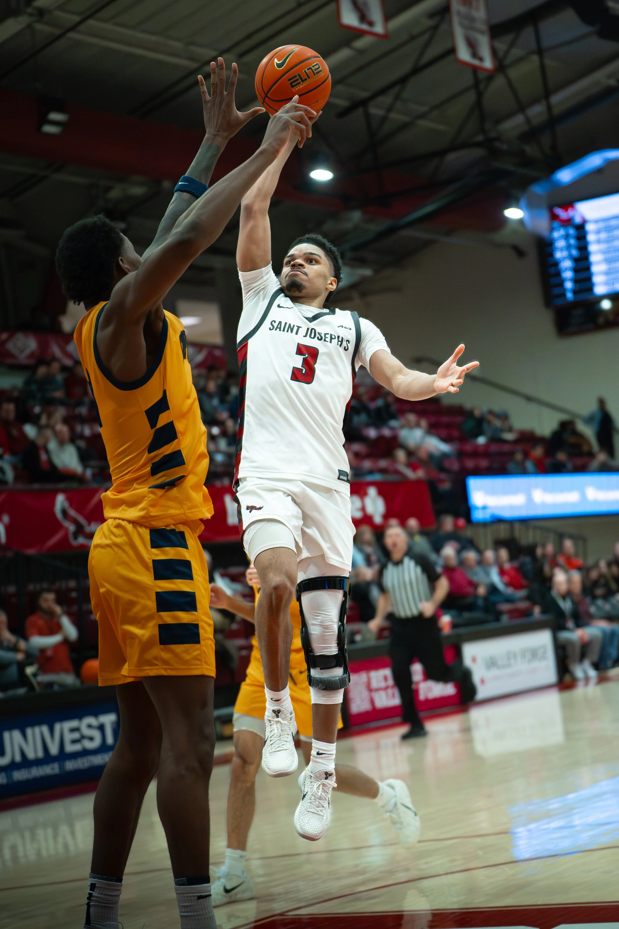 A basketball player from Saint Joseph's, wearing jersey number 3, jumps to make a shot while an opposing player in a yellow uniform attempts to block.