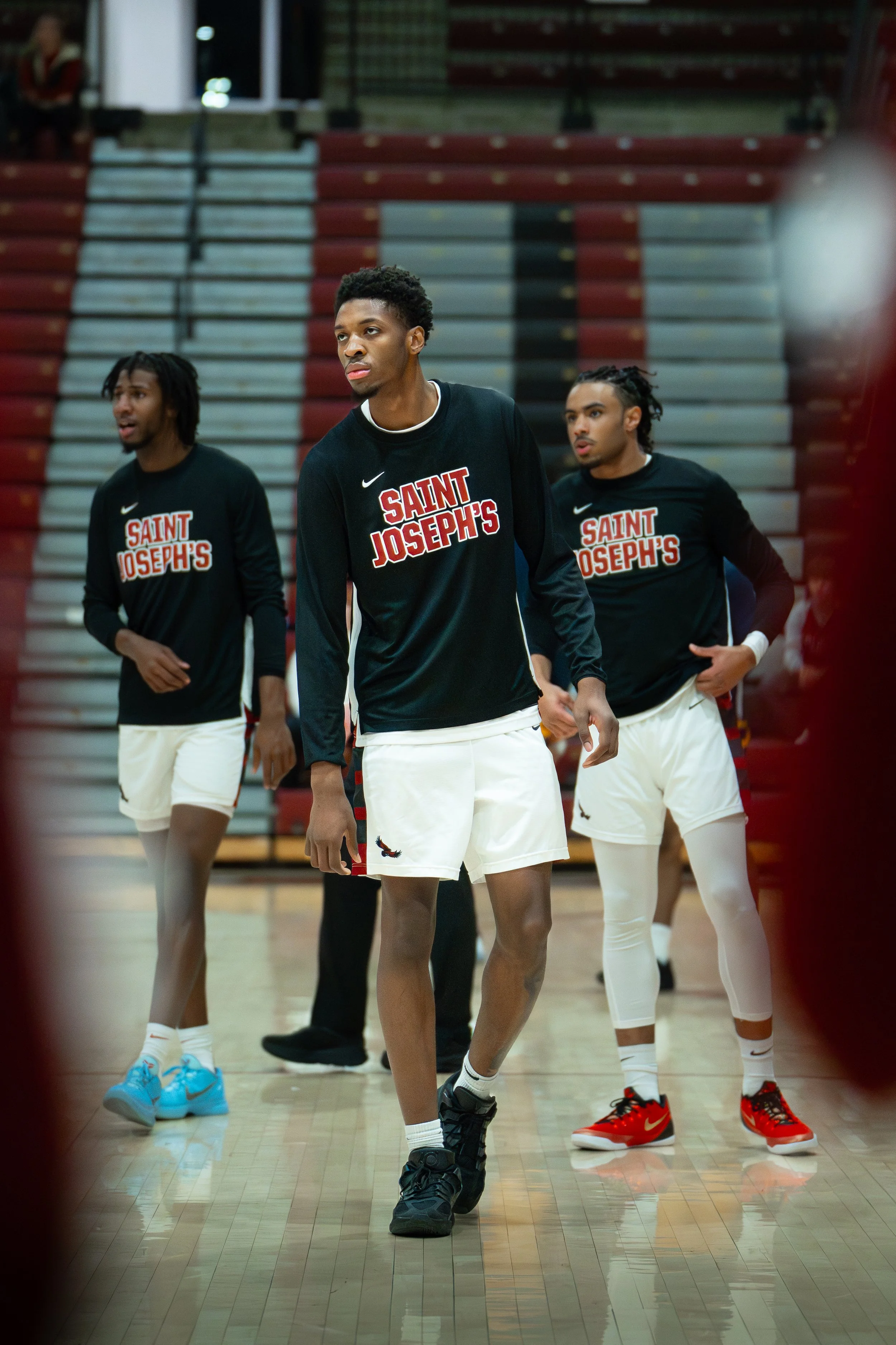 Three young men in basketball uniforms warming up on a court, wearing black and white Saint Joseph's team shirts.
