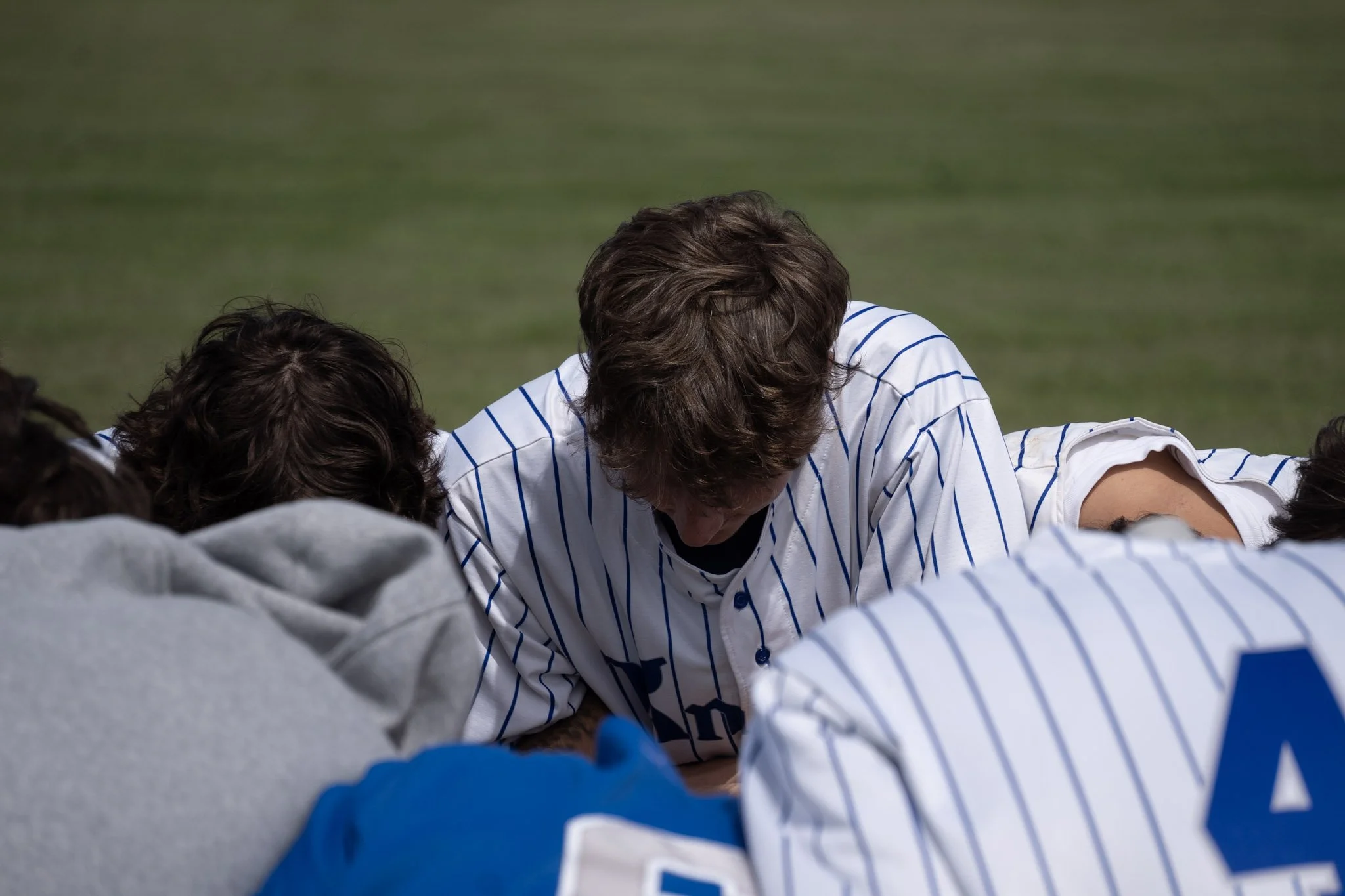 A group of people, including a man in a white and blue striped sports jersey, gather closely together with their heads bowed on a grassy field.