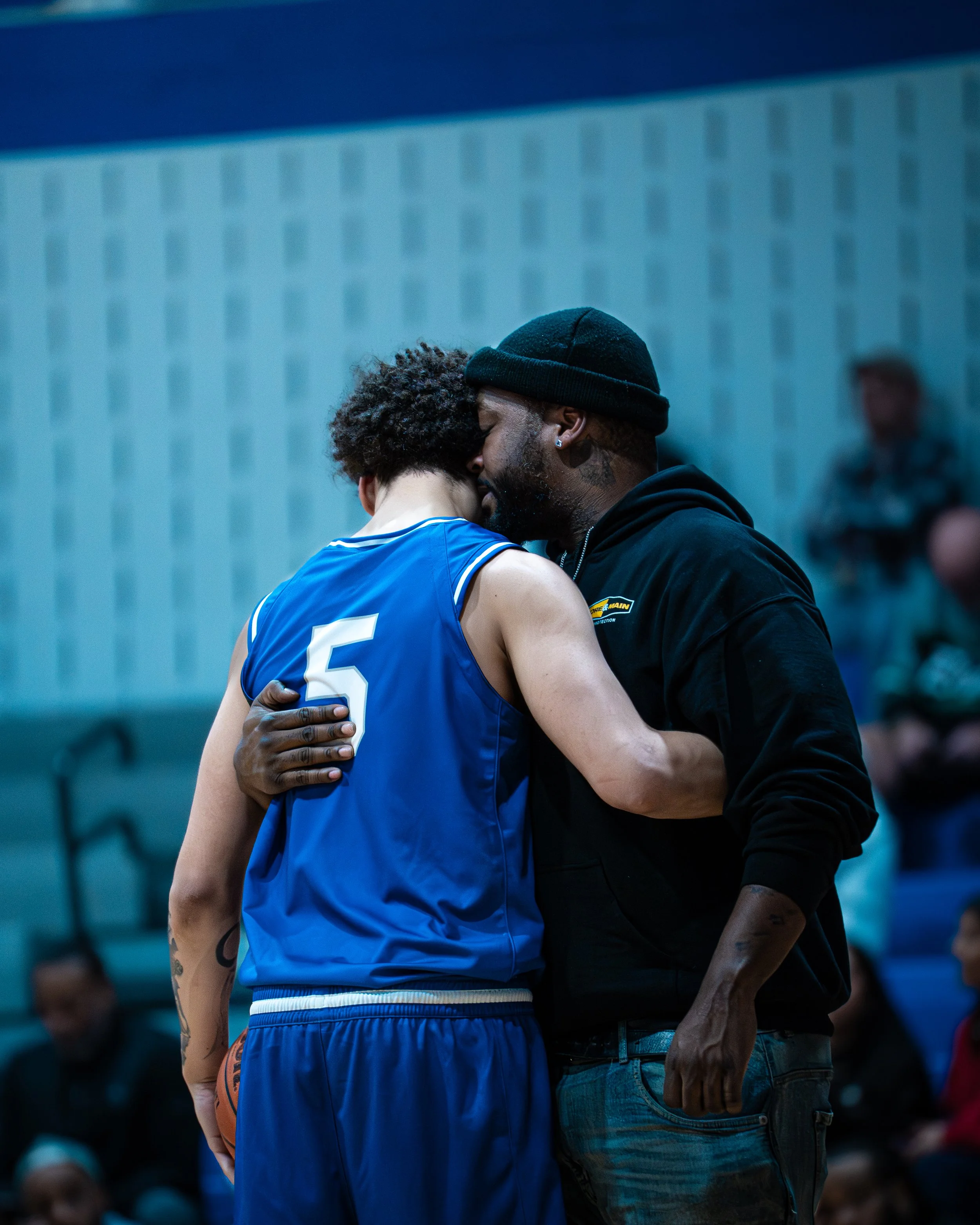 A basketball player in a blue jersey embraces a man in a black hoodie in a heartfelt moment during a game.