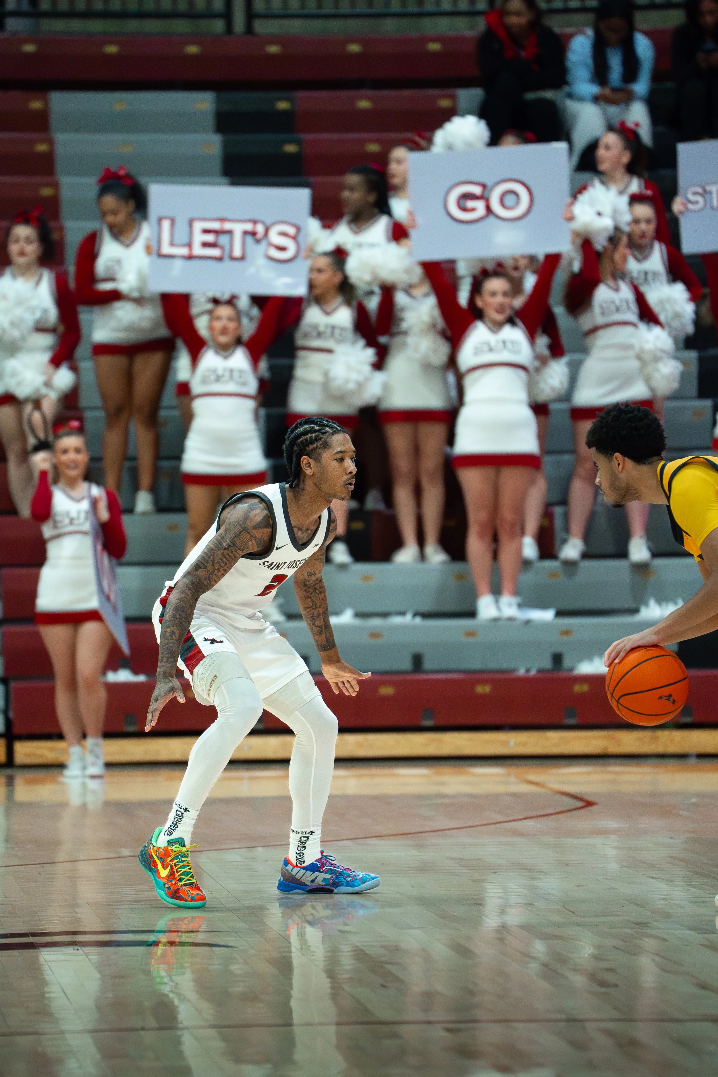A basketball game with a player in a white uniform and colorful shoes preparing to guard an opponent in a yellow jersey holding the basketball. In the background, cheerleaders hold signs that say "LET'S GO" and are wearing red and white uniforms.