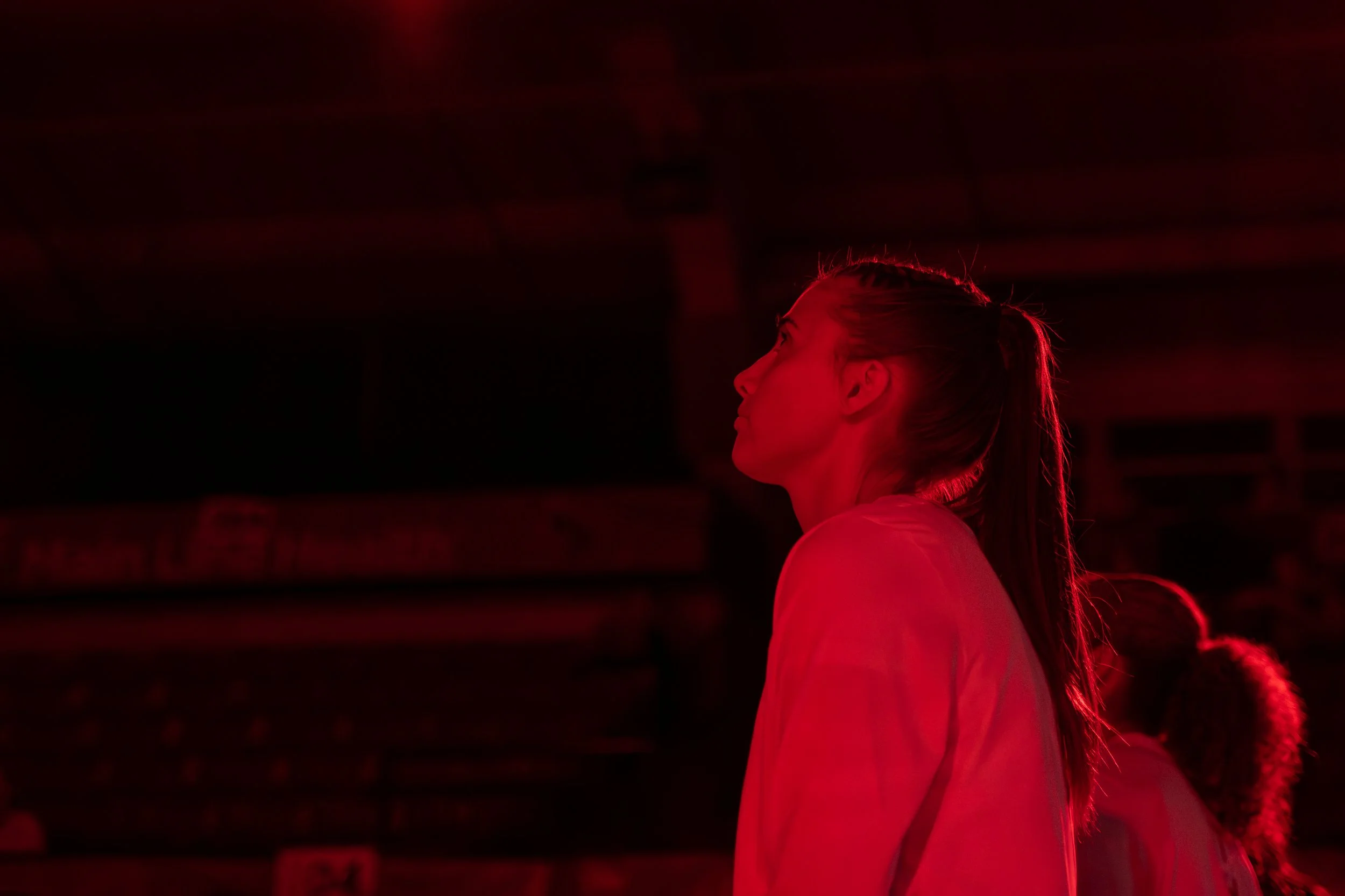 Side profile of a woman in a sports jacket illuminated by red lighting, with other individuals behind her, in an indoor sports arena.