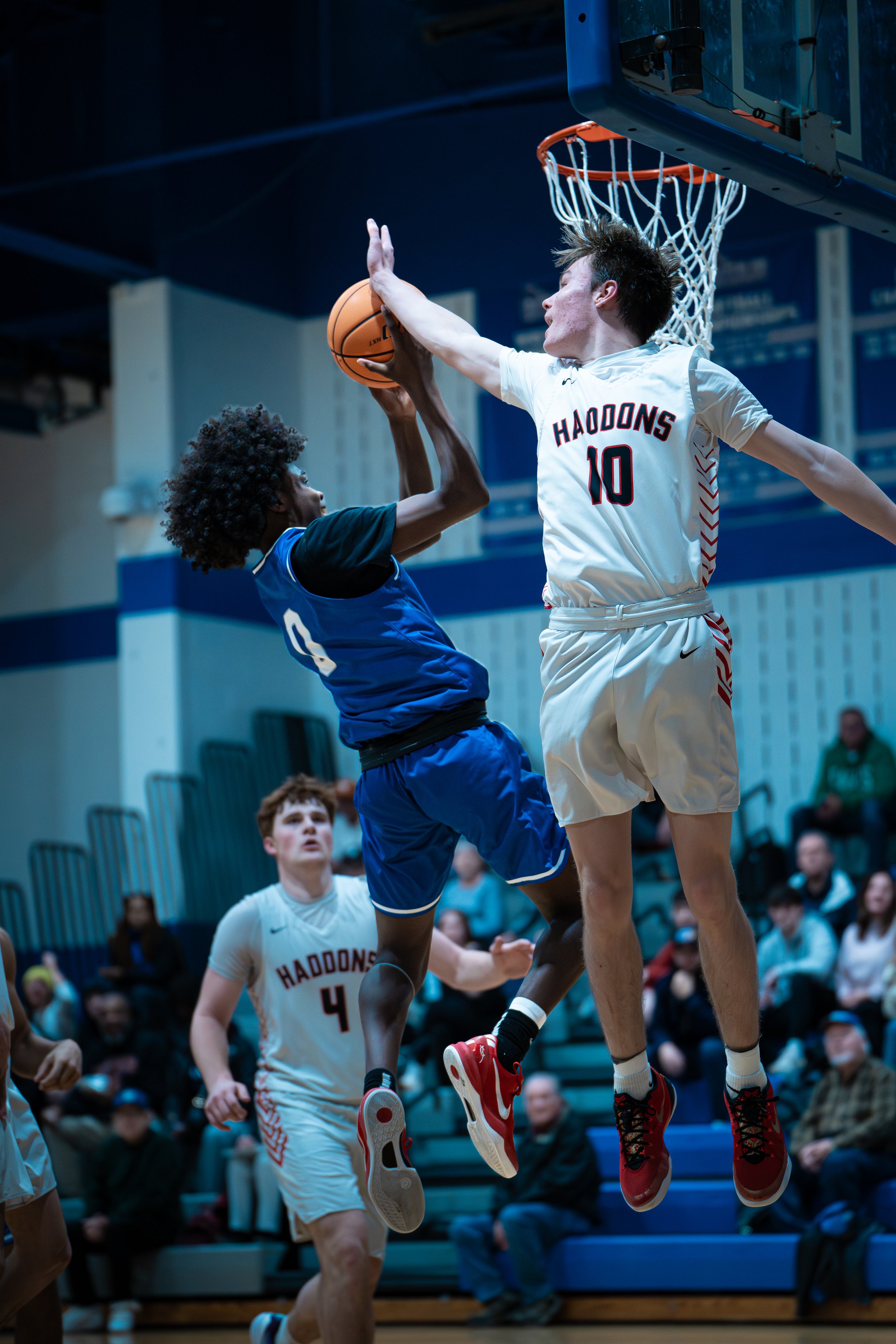 Two basketball players compete at the basket, with the player in blue jumping to block the shot of the player in white, while other players and spectators watch in the gym.