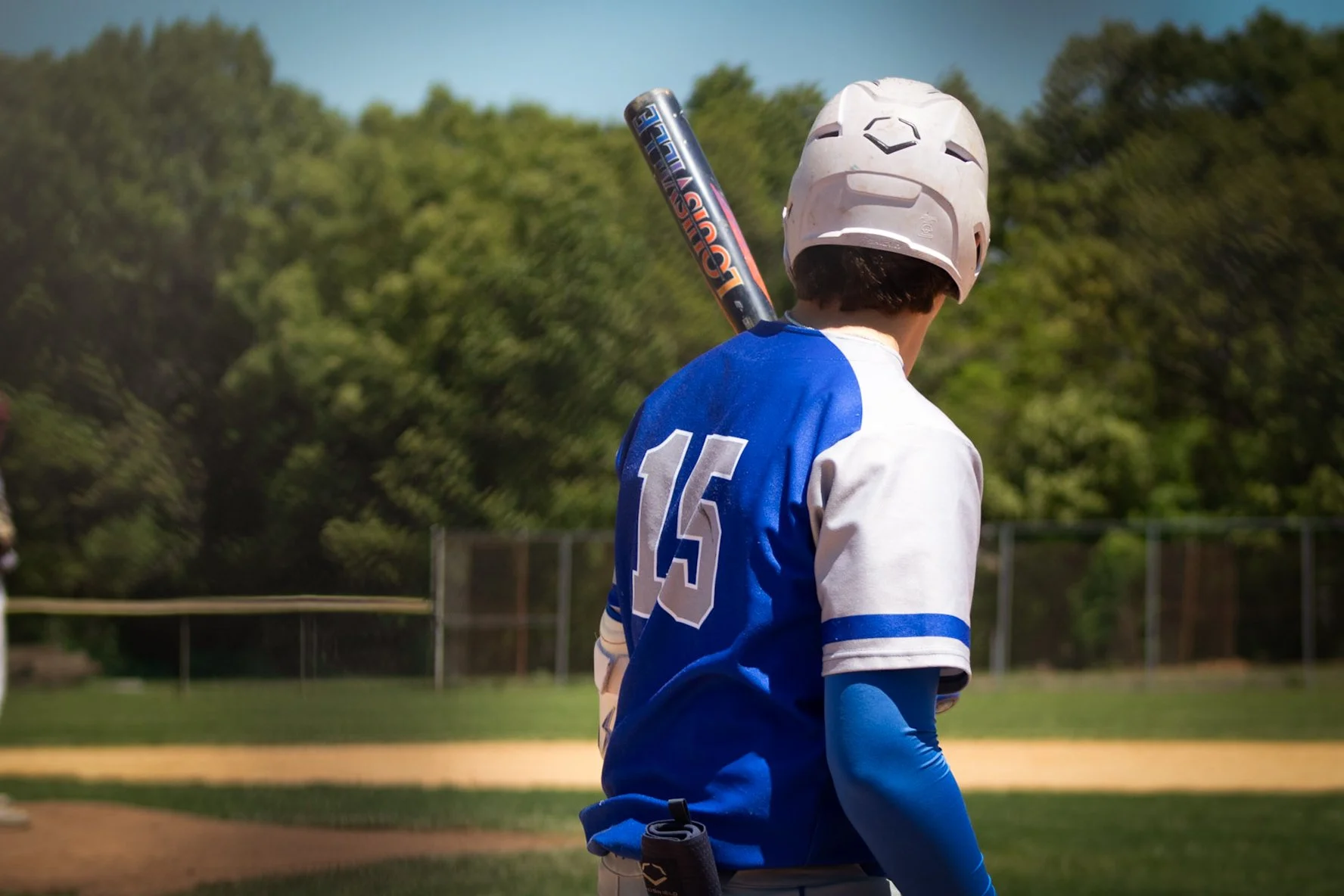 A young baseball player stands on the field holding a bat over his shoulder, wearing a helmet and a blue and white uniform with the number 15.