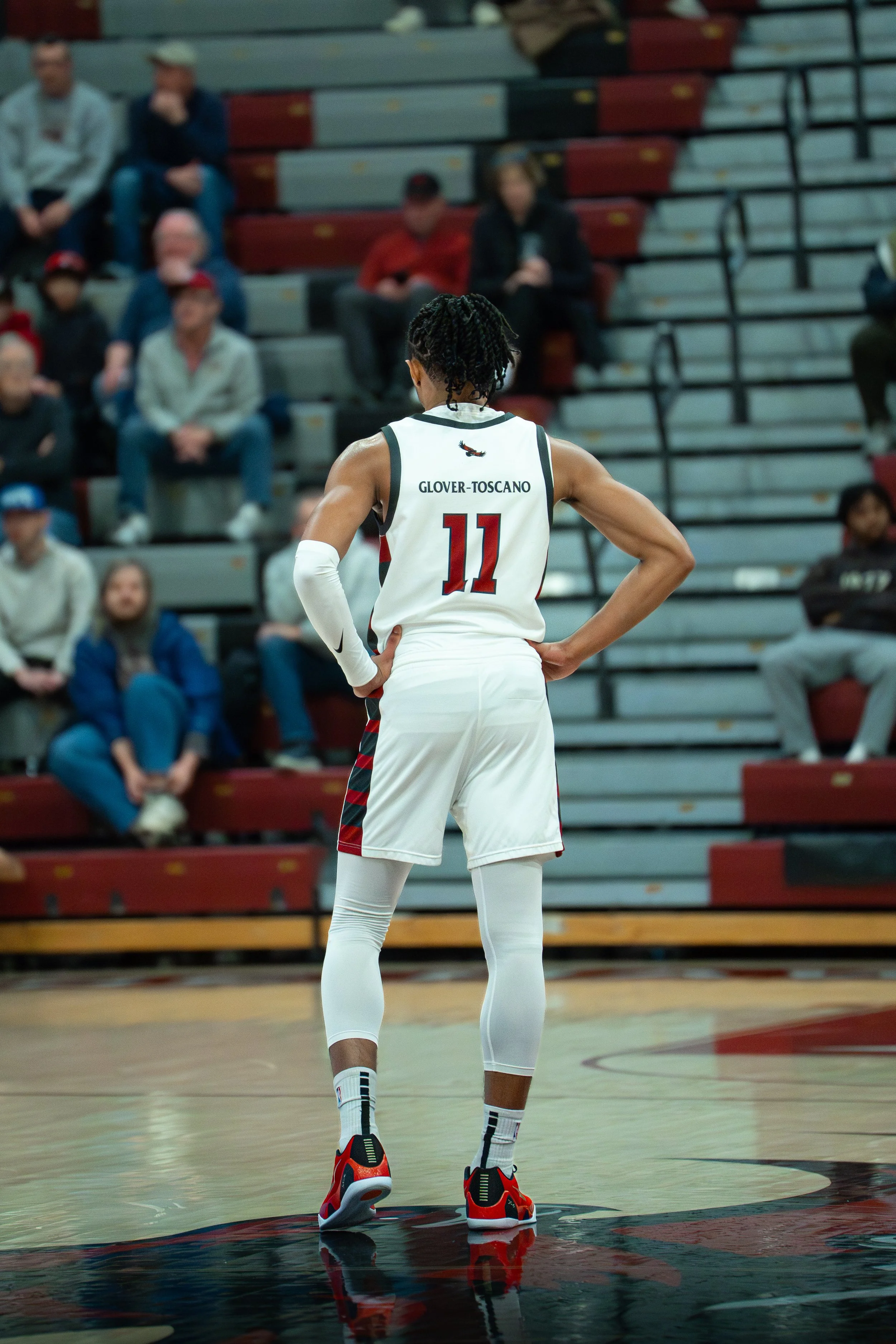 A female basketball player with jersey number 11 stands on a basketball court with her back to the camera, hands on her hips, in front of an audience seated in bleachers.