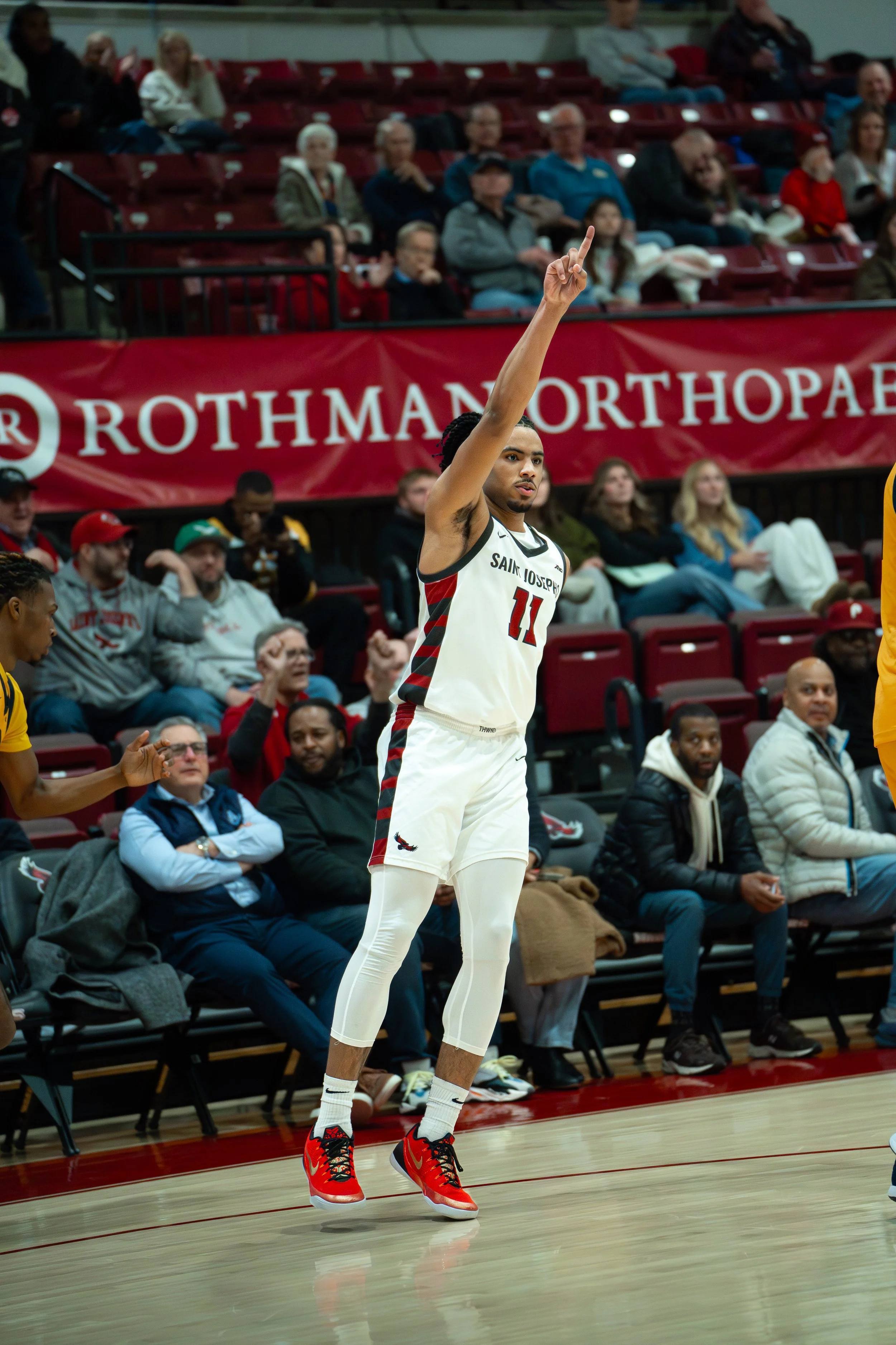 A basketball player in a white jersey with the number 11 and 'Saint Joseph' written on it, jumping on the court with one arm raised.
