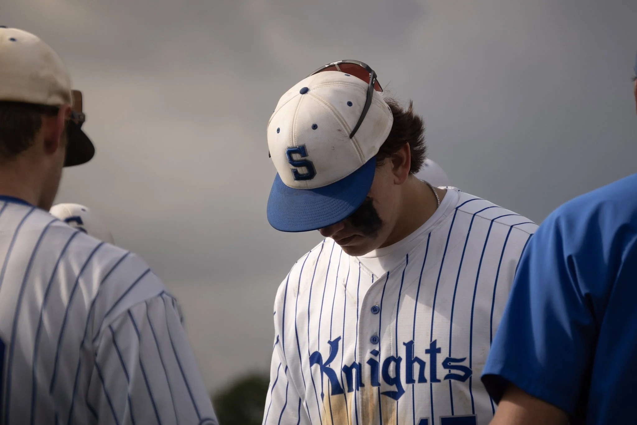 Baseball player wearing a pinstripe uniform and a cap with a letter 'S' on it, surrounded by teammates, looking downward during a pre-game ceremony or prayer.
