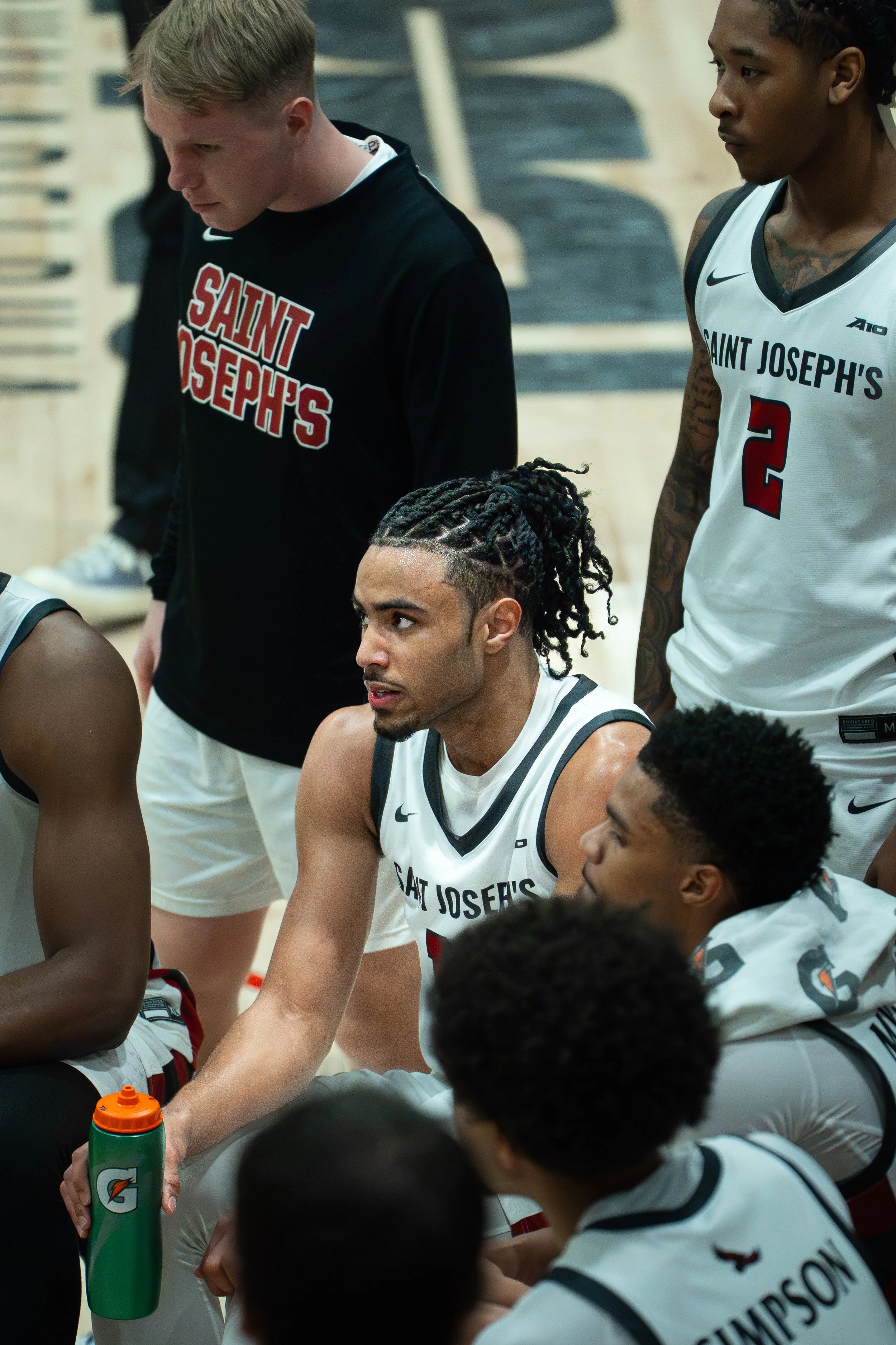 Basketball players and a staff member from Saint Joseph's team during a timeout or huddle, wearing white jerseys with black and red accents, with one player holding a Gatorade water bottle.