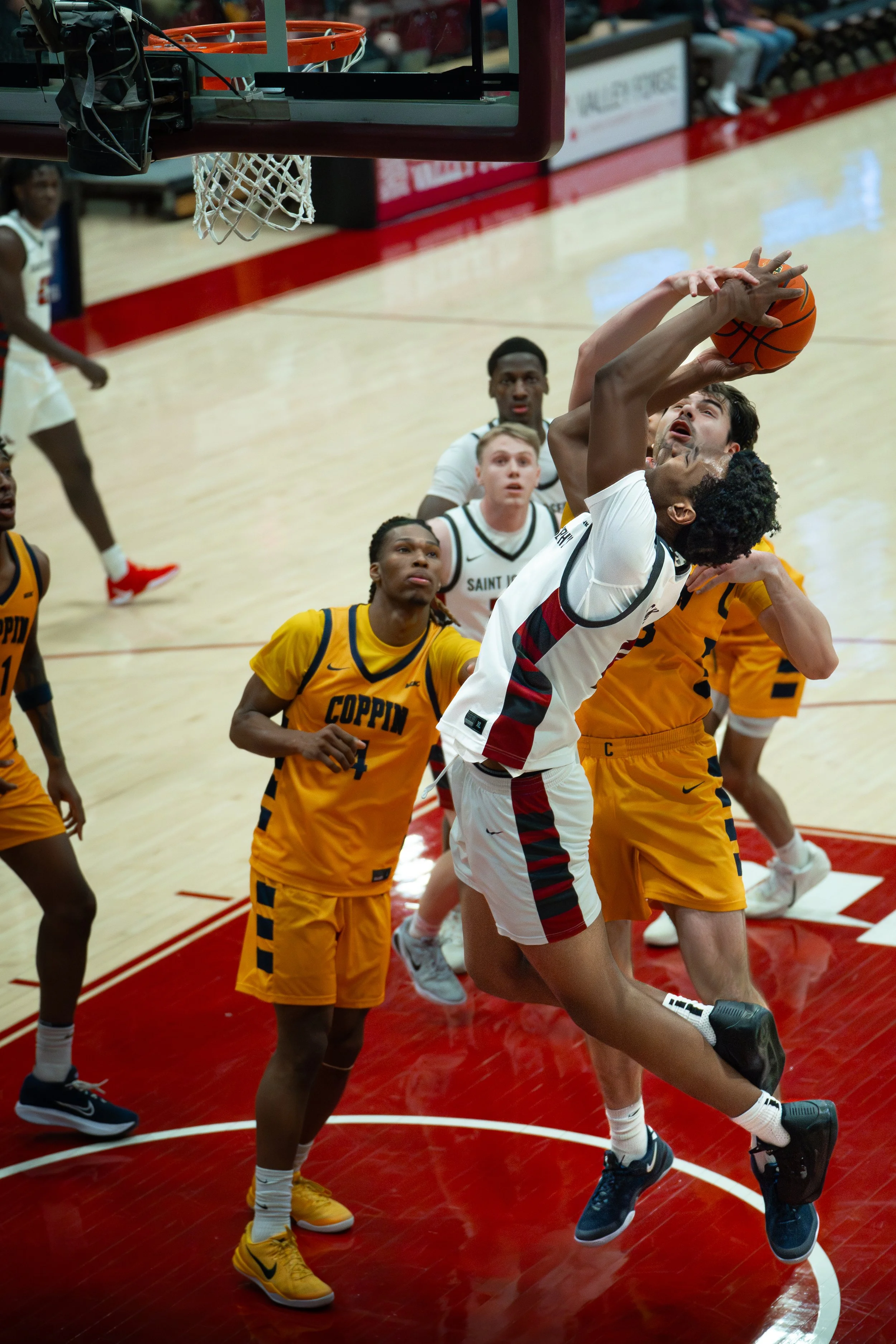 A basketball game with players jumping to shoot or block the ball near the basket, with players in yellow and white uniforms on the court.