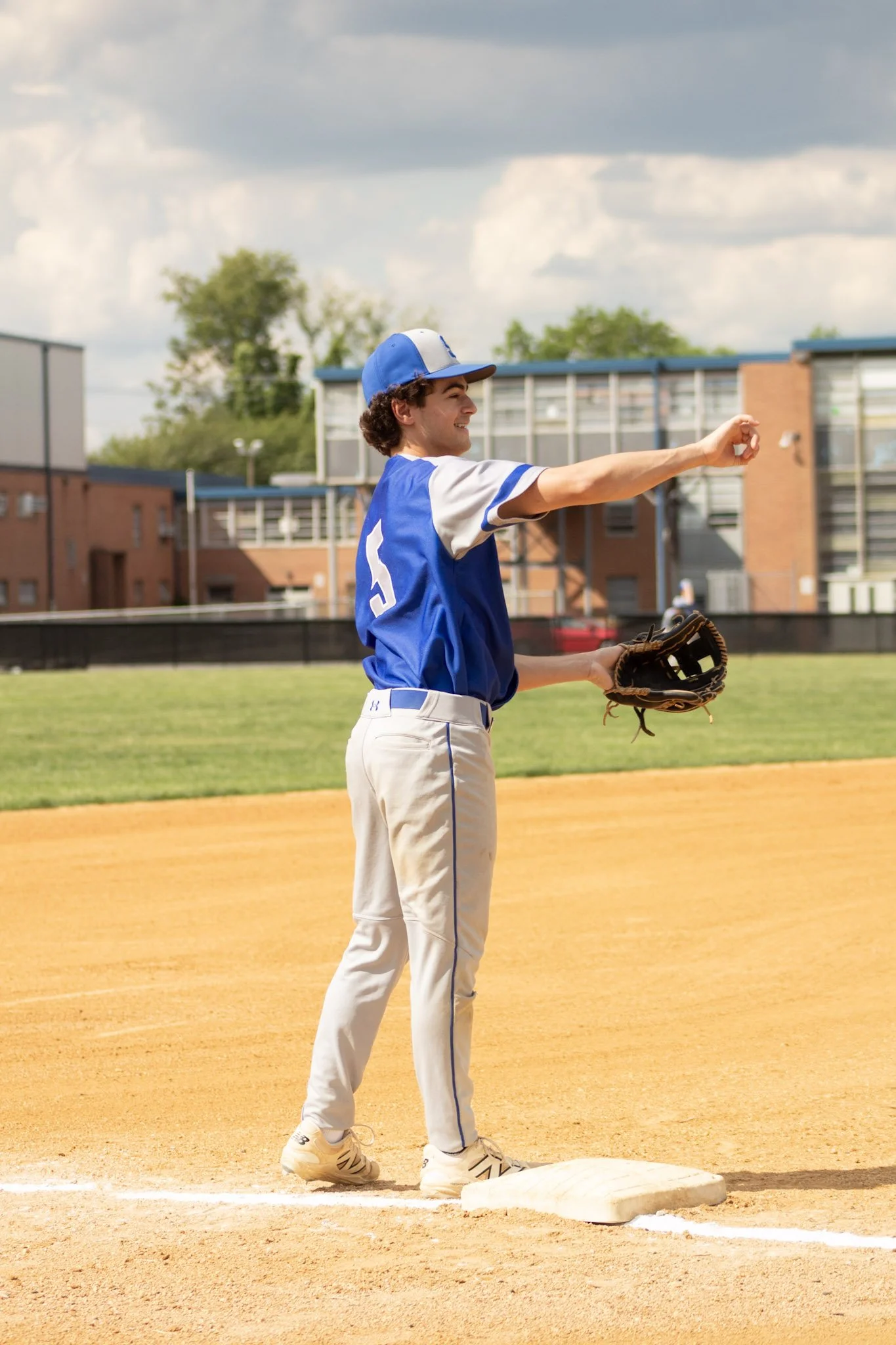 A young baseball player is standing on a base on a baseball field, throwing a baseball with a happy expression. He is wearing a blue and white uniform with the number 5 on his back, a matching cap, and a baseball glove on his left hand.