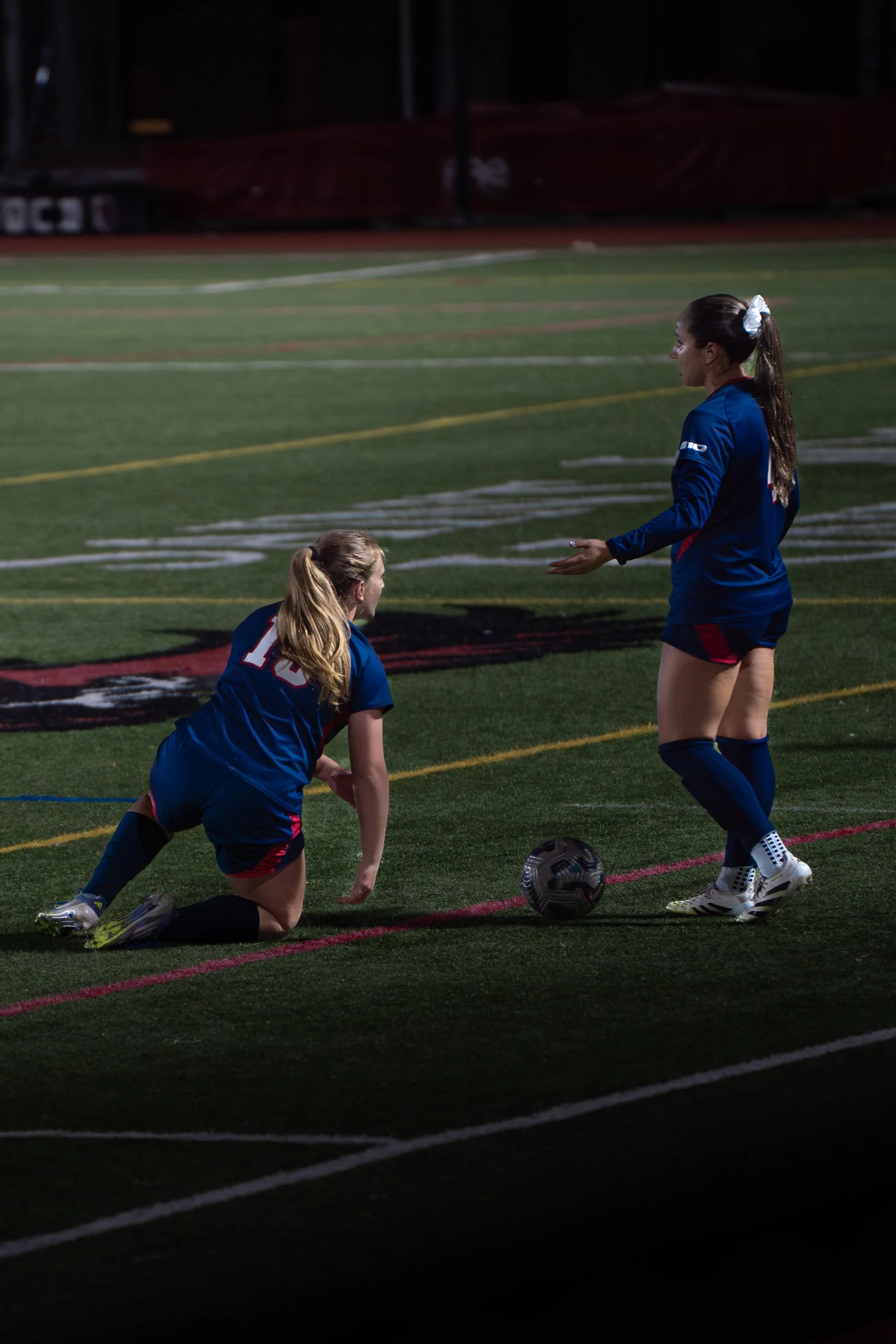 Two female soccer players in blue uniforms on a soccer field, one kneeling and the other standing, with a soccer ball near her foot, during a game at night.