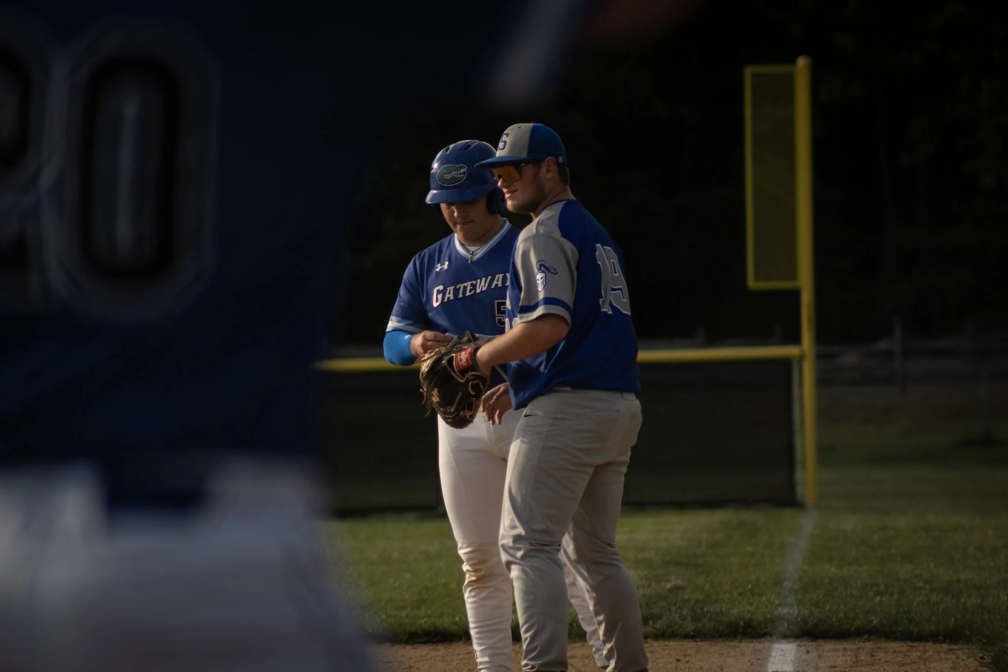 Two baseball players in blue uniforms on a field, one holding a baseball glove, with a yellow foul pole or scoreboard in the background.