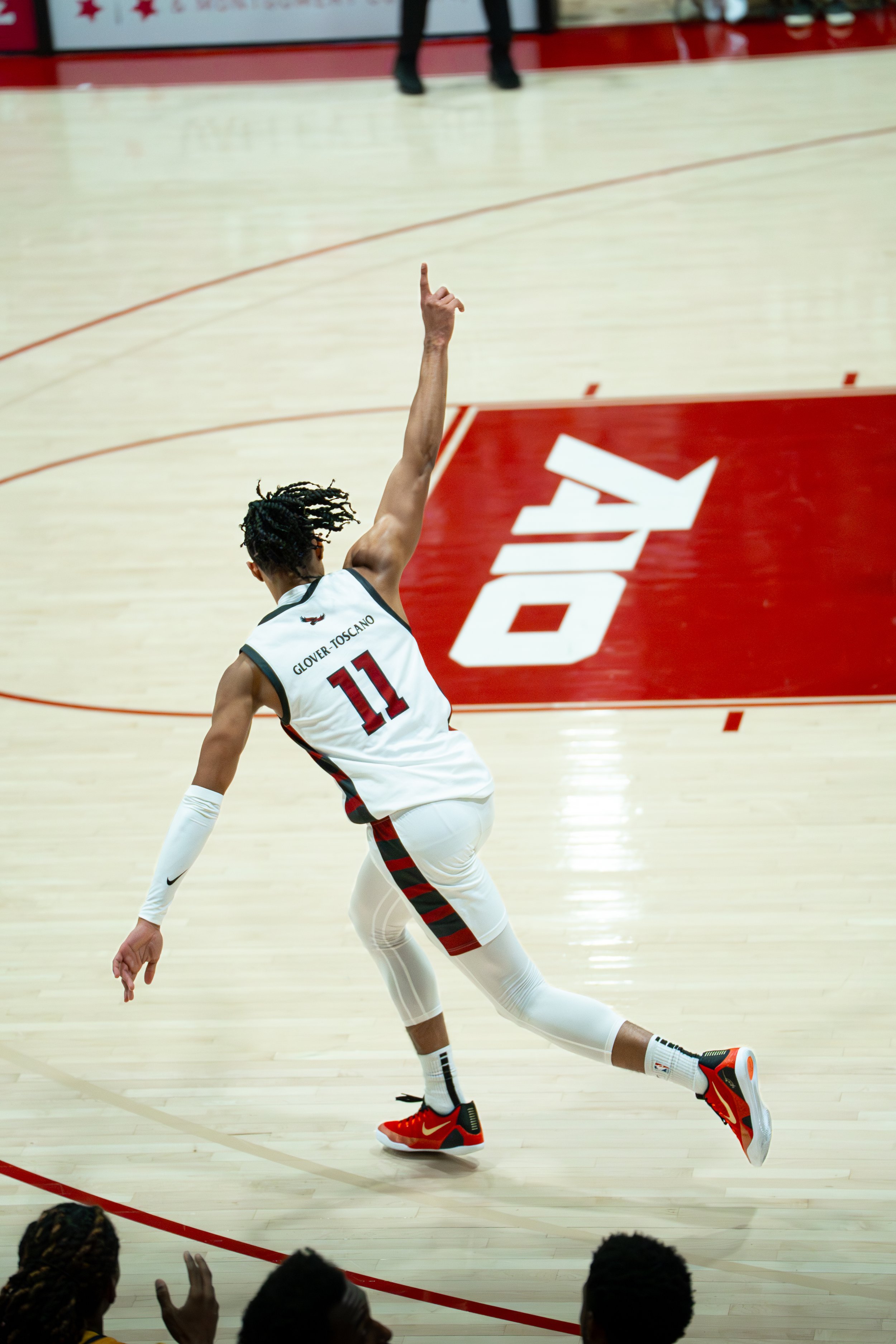 A basketball player wearing jersey number 11 with 'GLOVER TOSCANO' on the back, mid-action on the court, reaching up with one arm, with a crowd and red team logo in the background.
