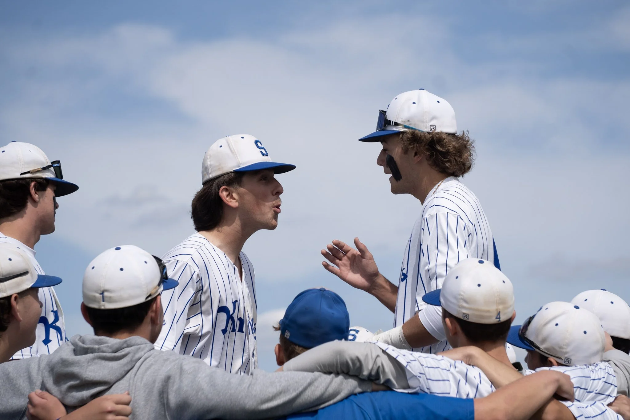 A group of youth baseball players in white and blue uniforms and caps, gathered around their coach who is speaking to them outdoors against a partly cloudy sky.