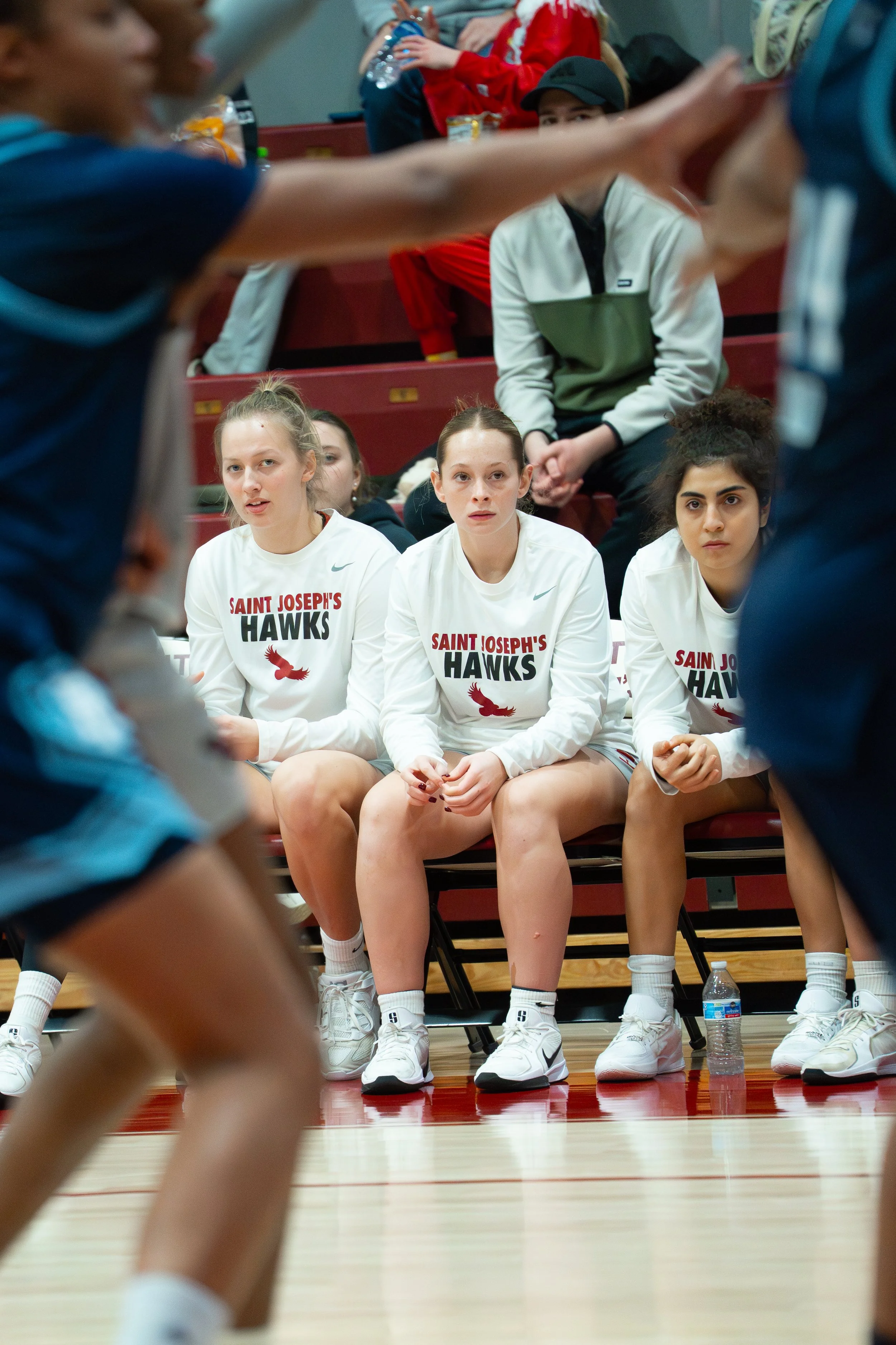 Girls' basketball team wearing white jerseys with 'Saint Joseph's Hawks' sitting on the sidelines of a game, watching players on the court.