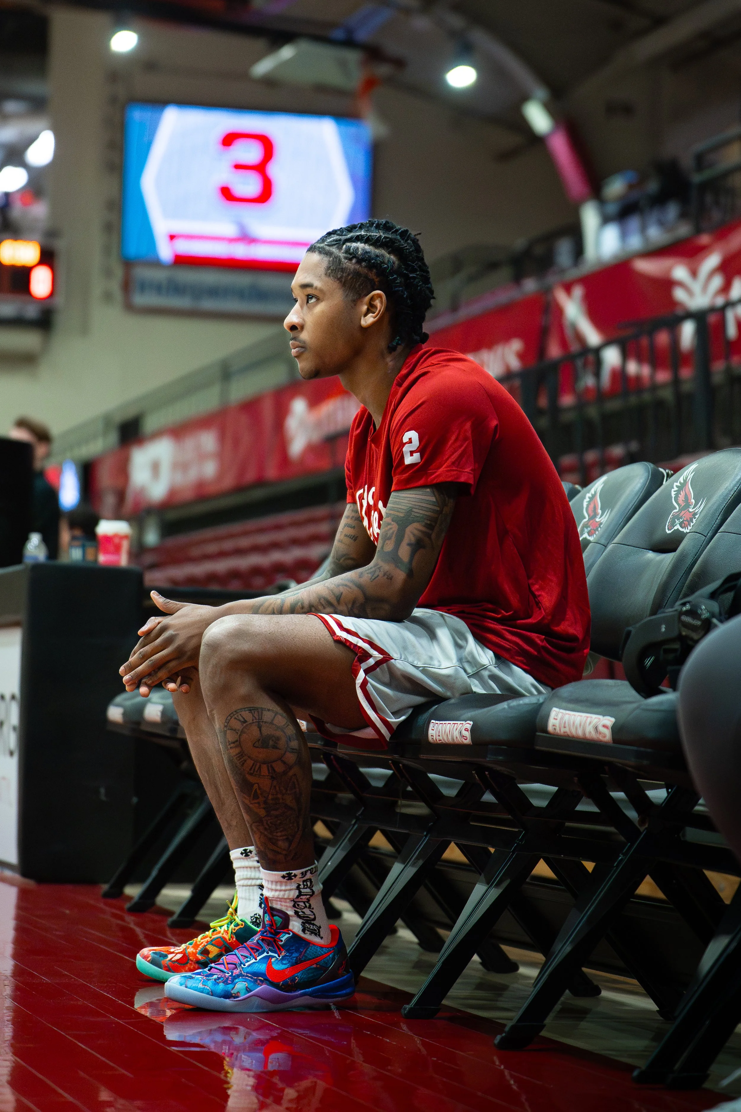 A young male basketball player with tattoos on his arms and leg, sitting on a bench in a sports arena, wearing a red jersey, colorful Nike sneakers, and white shorts with red trim.