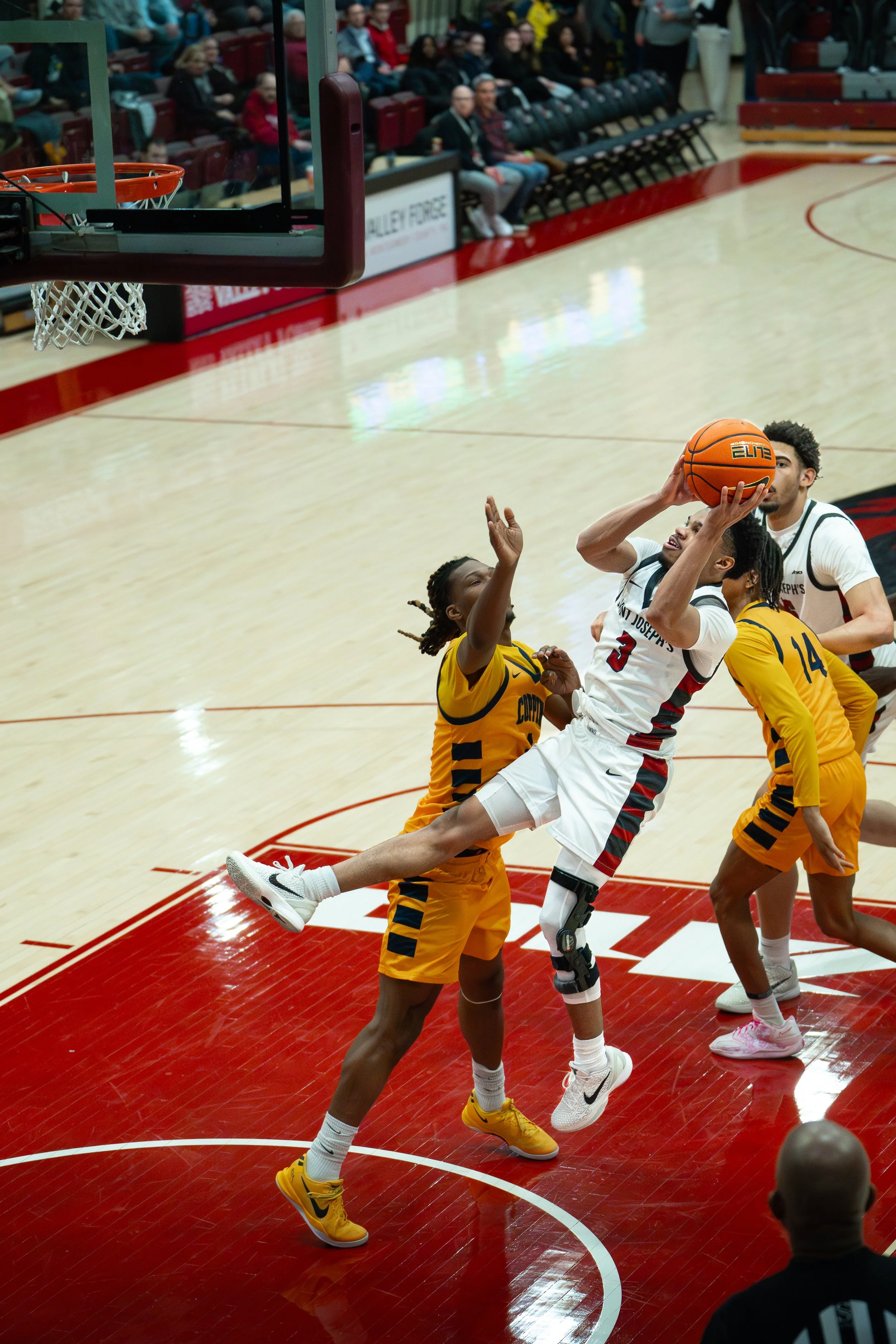 A women's basketball game with players in yellow and white uniforms. One player in white is jumping to shoot the basketball, while players in yellow defend. Spectators are visible sitting in the background.