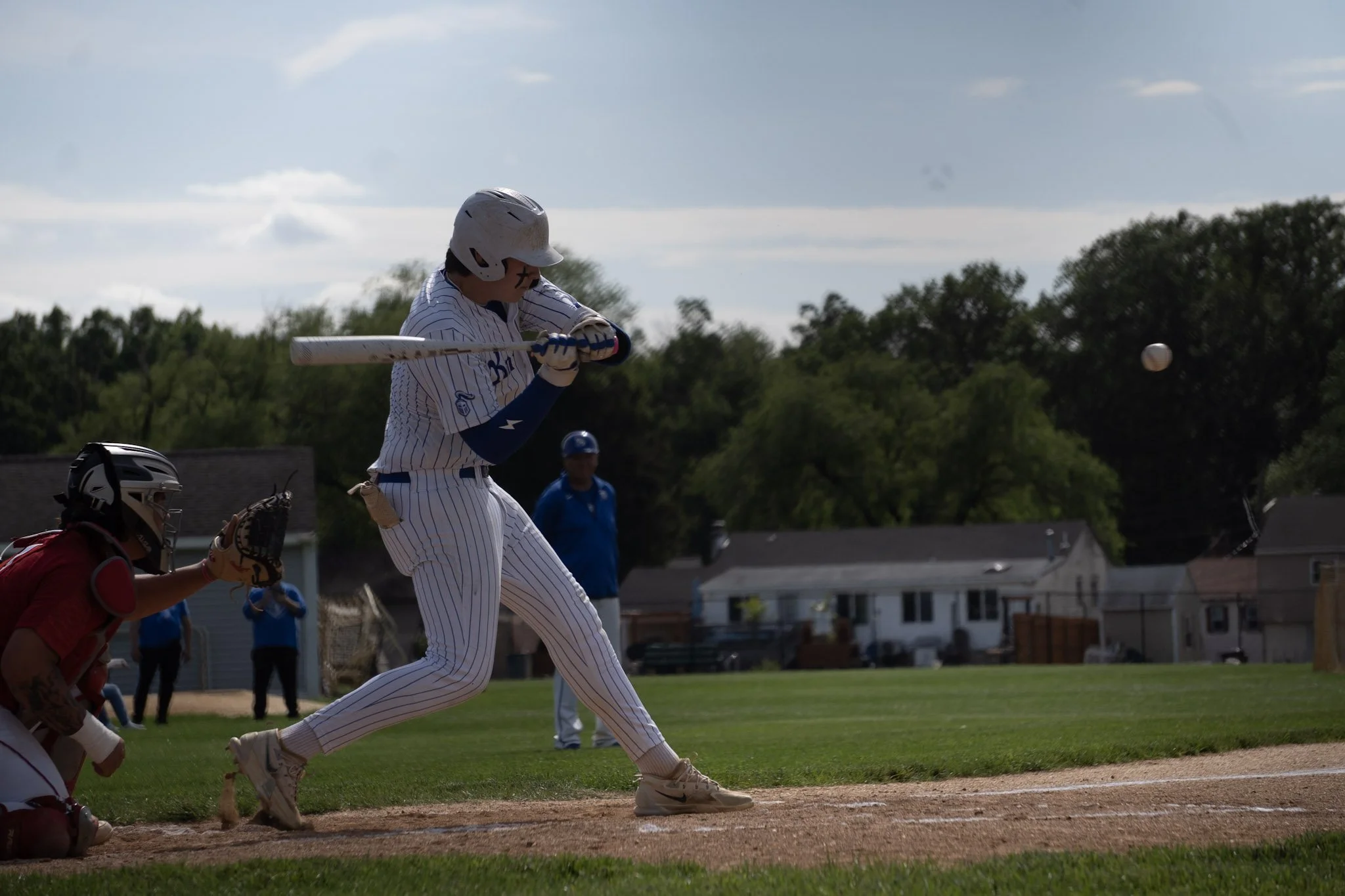 A baseball player in a white pinstripe uniform and helmet swings a bat at a ball during a game on a sunny day. The catcher in red gear and the umpire are behind him, with a grassy field and residential houses in the background.